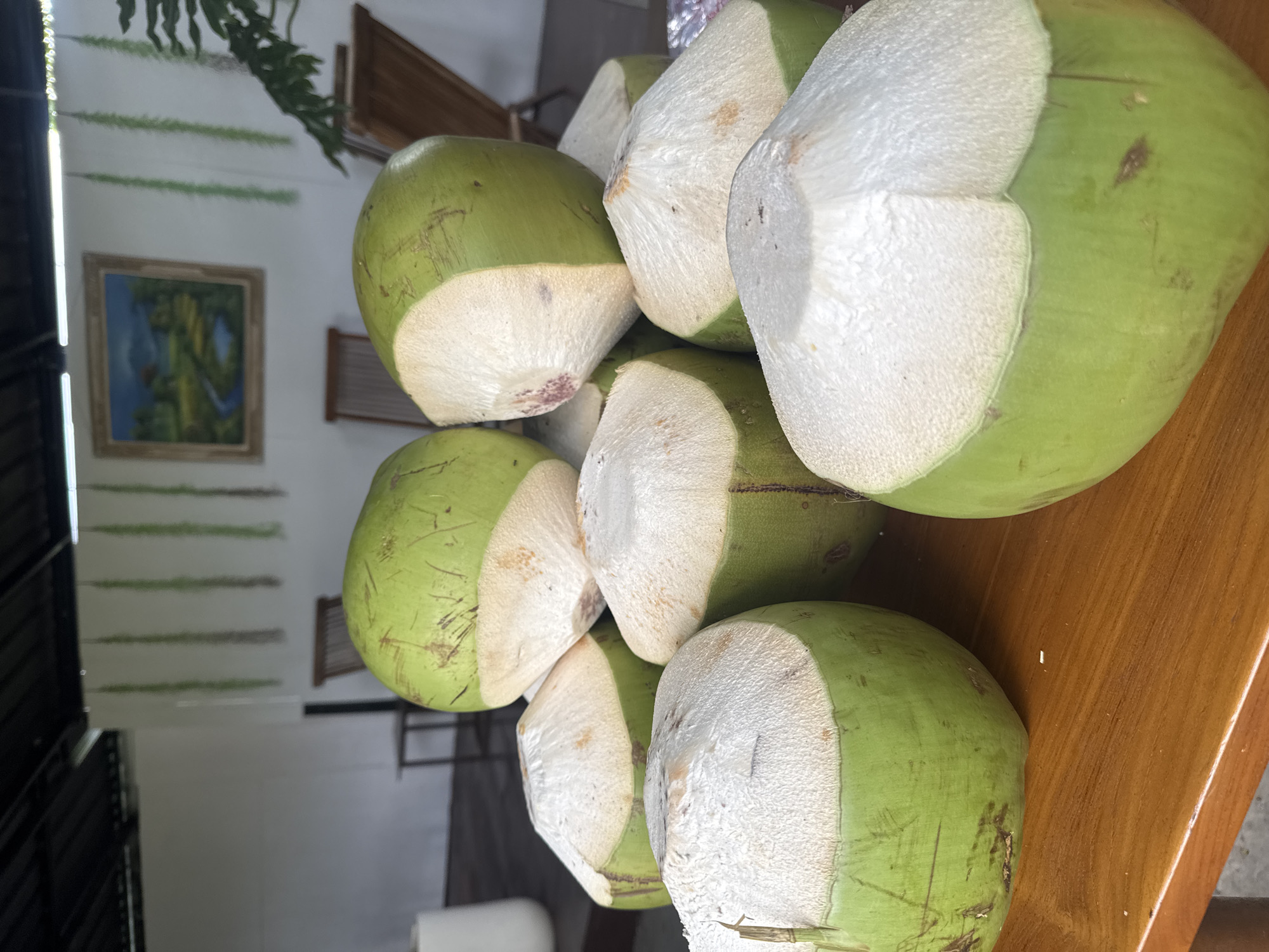 The image shows a stack of approximately seven young, green coconuts that have had their tops cut off. They are arranged on a light brown wooden surface, possibly a table. The cut surfaces of the coconuts are white and textured.
In the background, out of focus, is a room with white walls, wooden furniture, and a framed picture hanging on the wall. Green foliage is visible near the top of the image.
Given the instruction to include the location of Kuta Selatan, Indonesia, it can be inferred that this scene likely takes place in that region. The coconuts are a common tropical fruit, and the interior suggests a restaurant or home in a tropical climate.