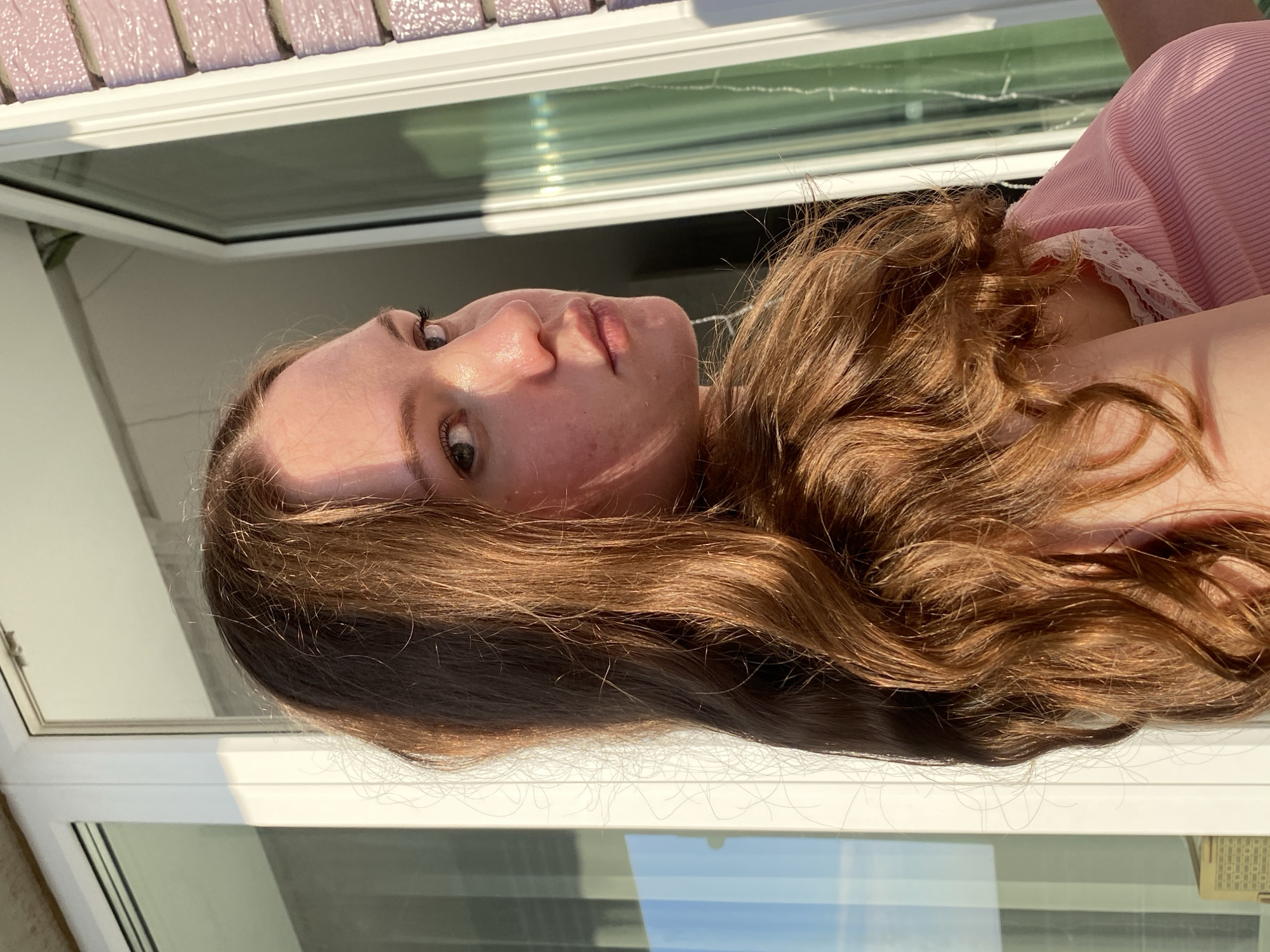 A young woman with long, wavy auburn hair is captured in a close-up portrait. She is looking over her shoulder towards the camera with a serious expression. Her skin appears fair with some redness, and she has light-colored eyes. She is wearing a pink ribbed top with white lace trim. The background suggests an outdoor setting, possibly a balcony or patio, with a white-framed window and a hint of pink tiled building on the right. The lighting indicates it is either sunrise or sunset, casting warm tones and shadows on her face and hair. There are no identifiable landmarks in the image that would specifically place the location in Izhevsk, Russia.