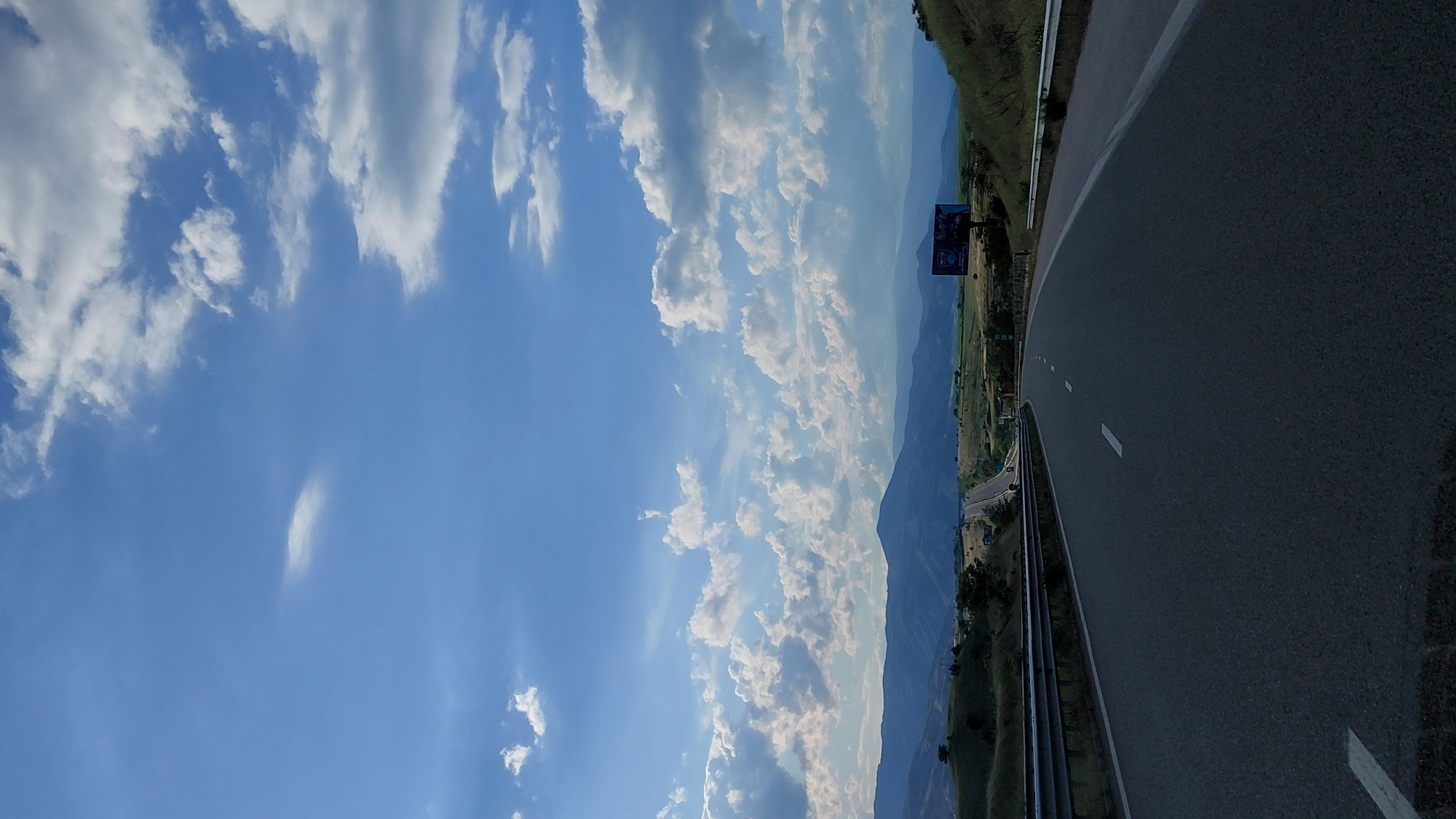 The image shows a road winding through a mountainous landscape under a partly cloudy sky. A billboard is visible along the roadside. The location is Unknown, Unknown.