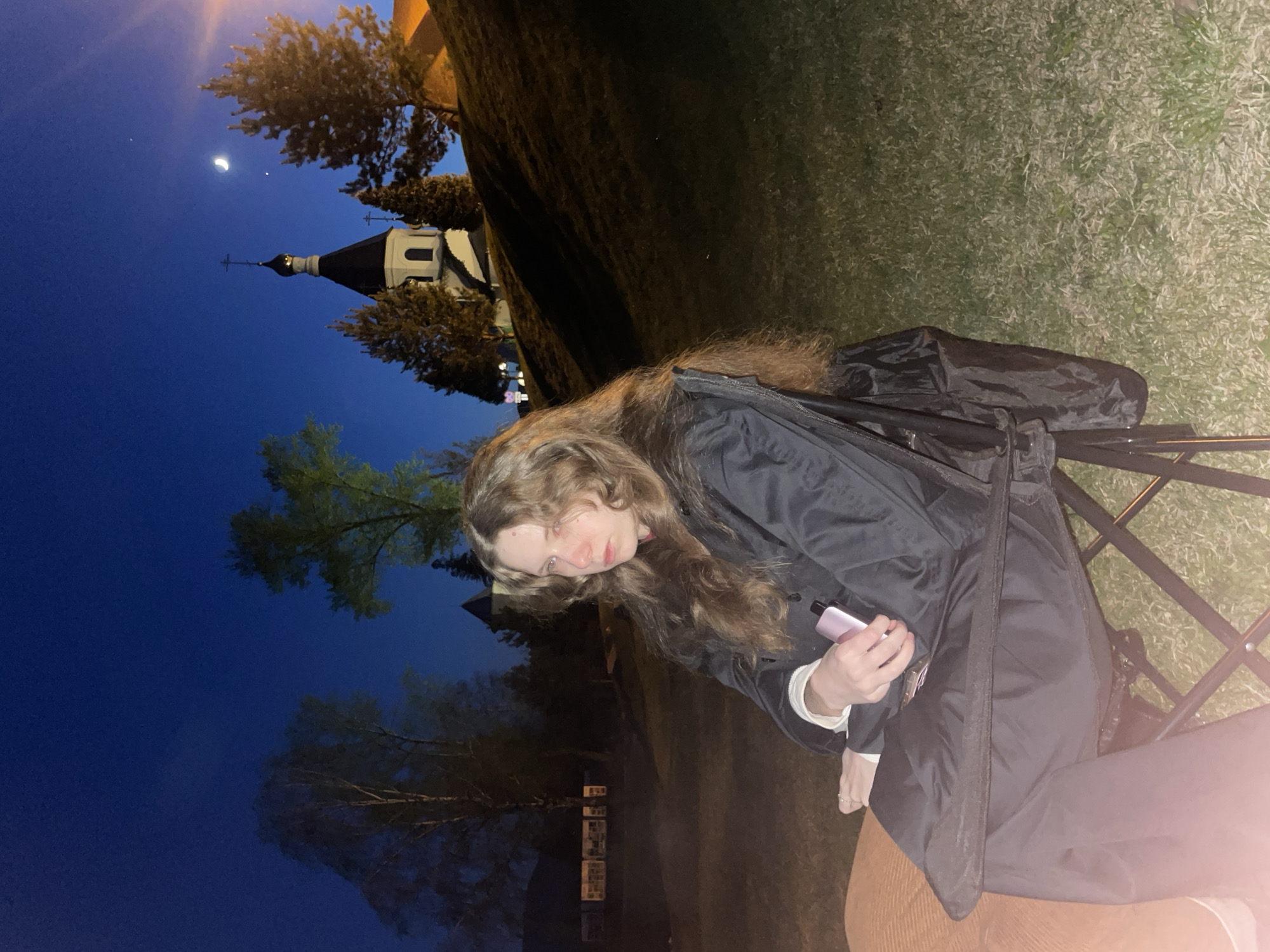 A woman with long brown hair sits on a folding chair, holding a pink object, possibly a vape, in her left hand. She wears a black jacket over a white long-sleeved shirt and brown pants. Behind her is a dark landscape with a church illuminated in the distance. The sky is a deep blue with a visible crescent moon. Trees are scattered around the scene, and the ground appears to be grassy. The location is likely in Krasnoyarsk, Russia, identified by the architecture of the church visible in the background.