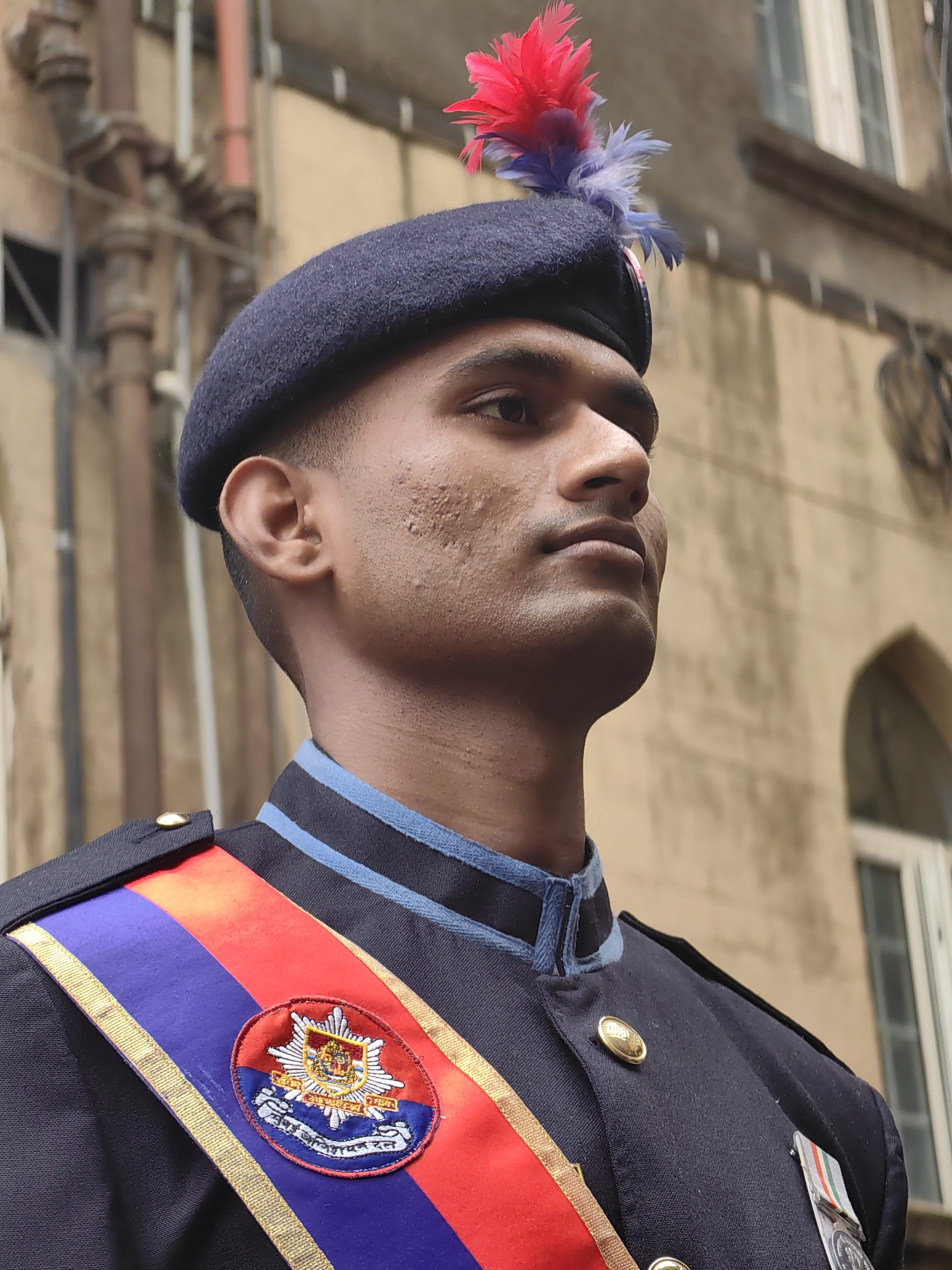 A close-up, medium shot captures a young man in uniform, standing outdoors. He wears a dark navy beret adorned with red and blue feathers. His uniform is dark navy with a light blue collar and trim. A sash with red and blue stripes, bordered by gold, crosses his chest, featuring a circular emblem. A medal with a tri-colored ribbon is pinned to his left breast. The man's face is turned to the right, looking upwards with a serious expression. His skin has some texture and appears lightly tanned. The background consists of a tan building with visible architectural details, including windows and what appears to be piping or conduit. The scene is consistent with a formal occasion, possibly a parade or ceremony, in a city environment.