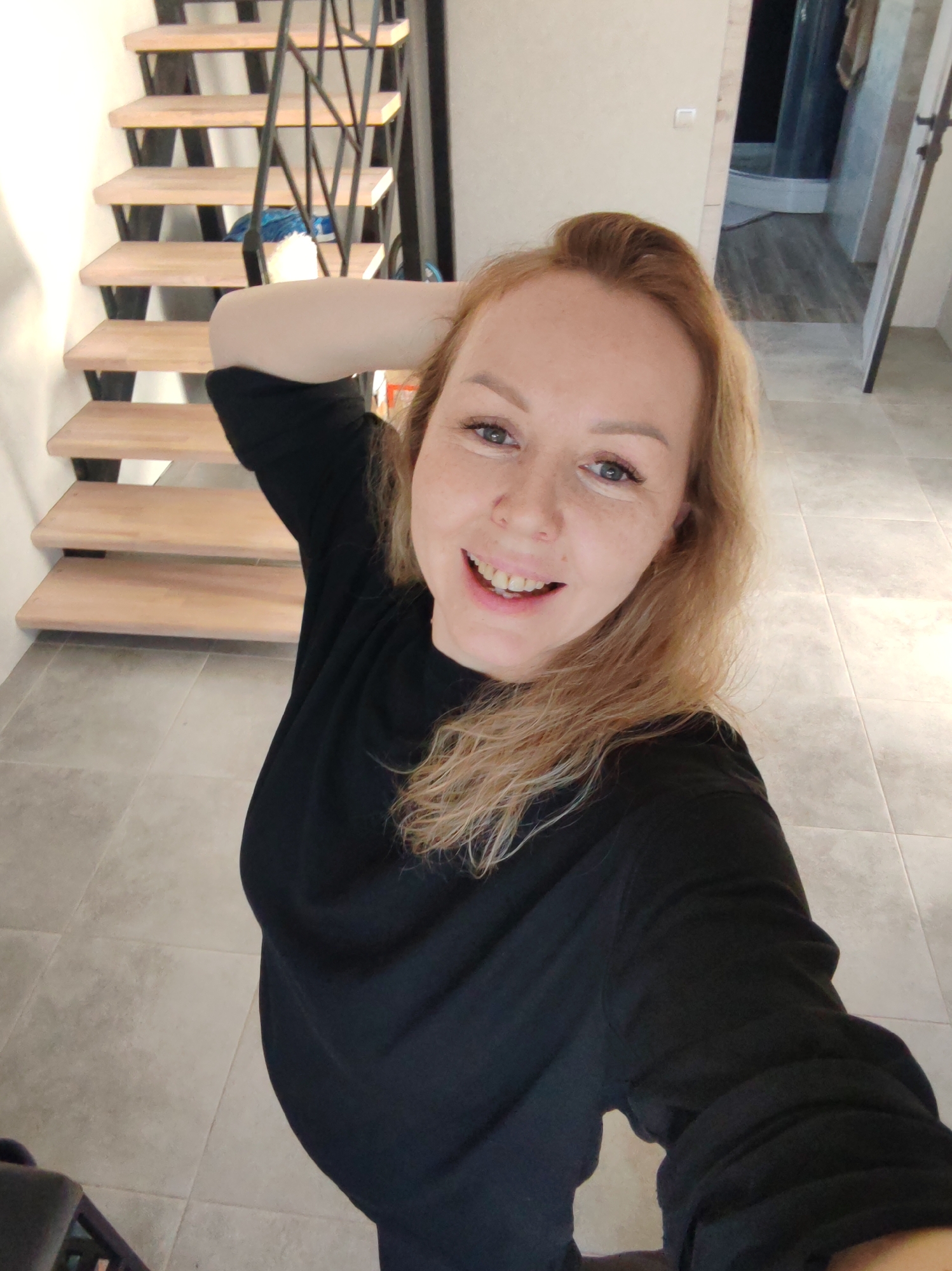 A woman with light blonde hair and freckles is taking a selfie indoors. She is wearing a black long-sleeved shirt. Her right arm is raised and her hand is behind her head. A staircase with wooden steps and a black metal railing is visible in the background. A doorway leading to what appears to be a bathroom is also visible in the background. The floor is covered in large gray tiles. The location is in an indoor space, likely a house or apartment. There is no indication of the location within the city of Chaikovsky, Russia.