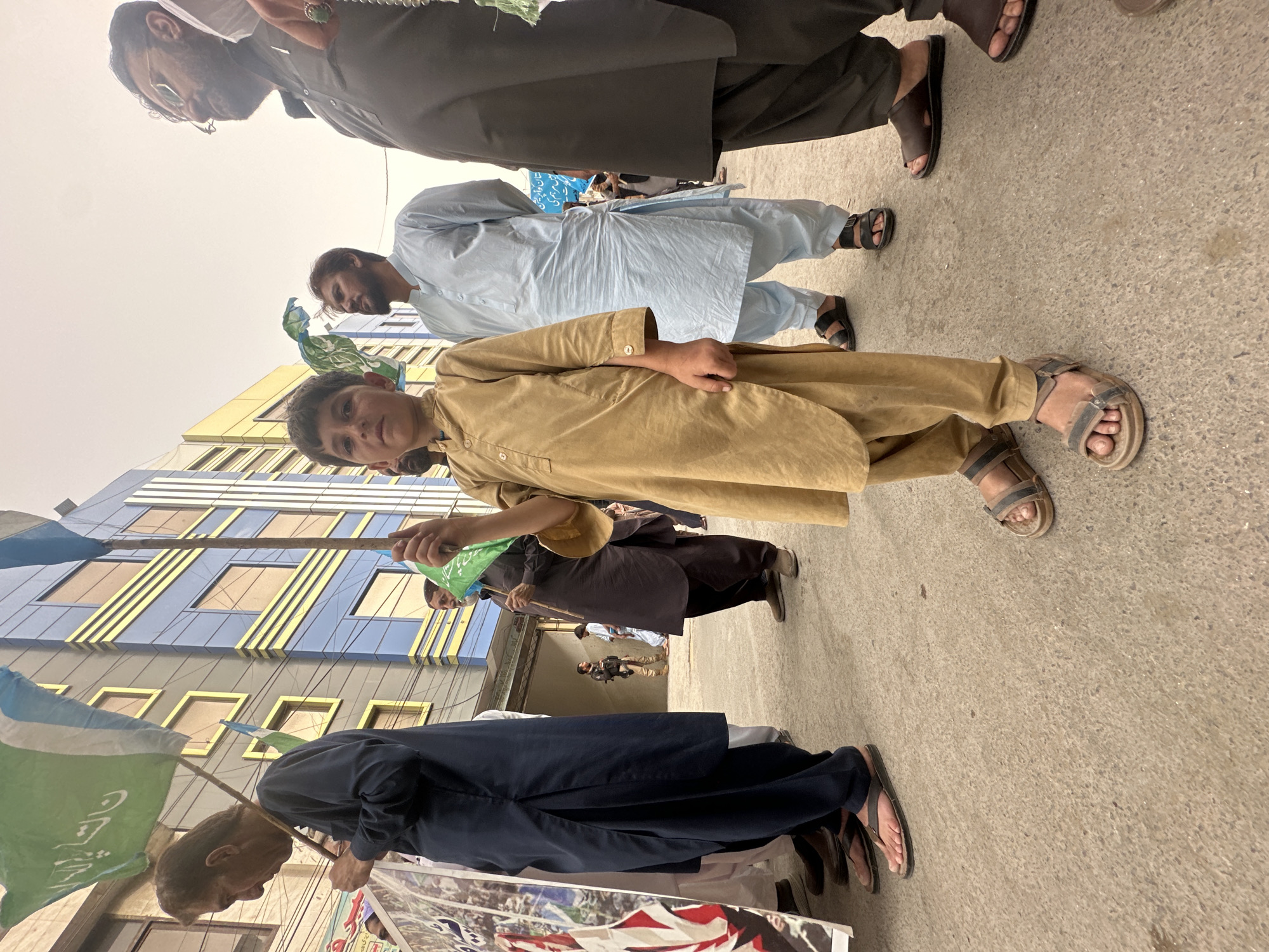 A low-angle, medium shot captures a group of individuals in Quetta City Tehsil, Pakistan, during what appears to be a procession or gathering. In the foreground, a young boy, possibly of Pashtun ethnicity, stands prominently. He wears a light brown shalwar kameez and sandals. The boy holds a green flag attached to a wooden pole, which he holds up with both hands. Behind him, a man in a light blue shalwar kameez stands with his hands clasped behind his back. To the boy's right, another man in a dark brown shalwar kameez is visible, also holding a green flag on a pole. Further to the right, the shoulder and head of a man wearing glasses and a dark outfit are seen.
In the background, a multi-story building with blue and yellow facade elements rises behind the people. Other individuals are dispersed throughout the scene, some holding flags or banners. The ground is paved with a rough, grey surface. The sky is overcast and pale. The overall atmosphere suggests a public event, possibly a religious or political demonstration, given the presence of flags and the group's formation. The image appears to be taken outdoors, on a street or in an open area within the city.