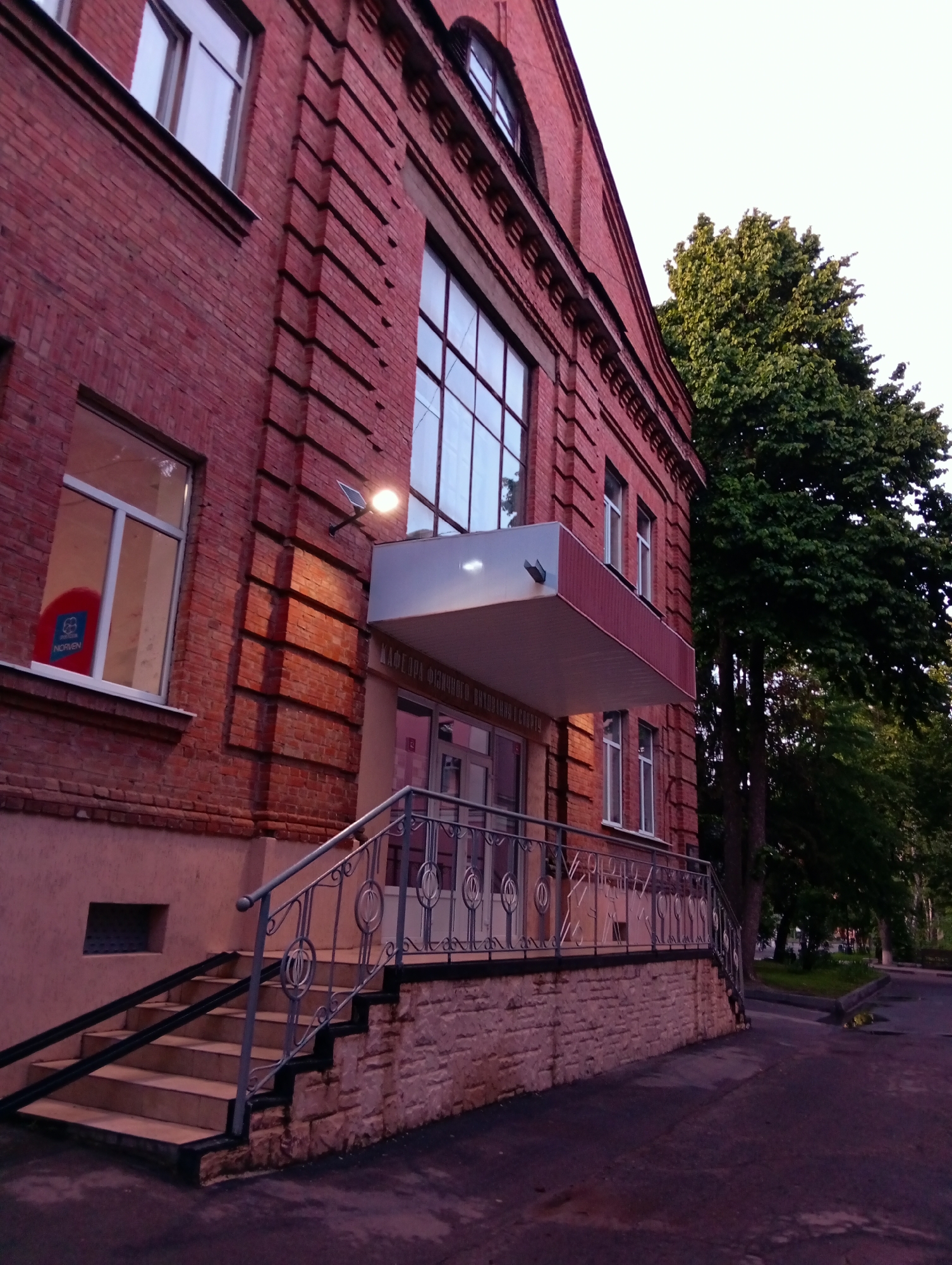 A low-angle shot captures a red brick building in Poltava, Ukraine. The building has a prominent facade with vertical brickwork extending from the window frames. A small balcony with a flat roof juts out above the entrance. Windows are visible on multiple floors, and a light fixture is positioned above the entrance. Steps lead up to the entrance, bordered by a decorative metal railing. The sidewalk runs along the building, and trees are visible on the right side of the frame. The lighting suggests late afternoon or early evening.