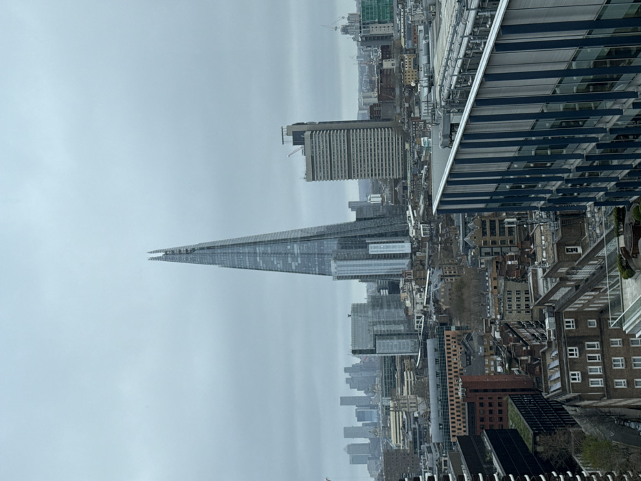 The image displays an elevated panoramic view of the London cityscape under a uniformly overcast, light grey sky. The prominent central feature is The Shard, a towering, glass-clad skyscraper with a distinct jagged, pointed apex, situated in the London Borough of Southwark. To the left, in the distant background, a cluster of modern high-rise buildings identifies the Canary Wharf skyline. The mid-ground reveals a dense urban tapestry composed of various architectural styles, including contemporary glass office blocks and numerous older, predominantly brick residential and commercial structures. To the immediate right of The Shard, a multi-story, light-colored block-like building features a construction crane on its roof. In the lower right foreground, the upper section and a rooftop terrace of a modern building, characterized by vertical blue and grey facade elements, are partially visible. No individuals are discernible within the frame. The scene presents a densely built-up metropolitan area within the United Kingdom.