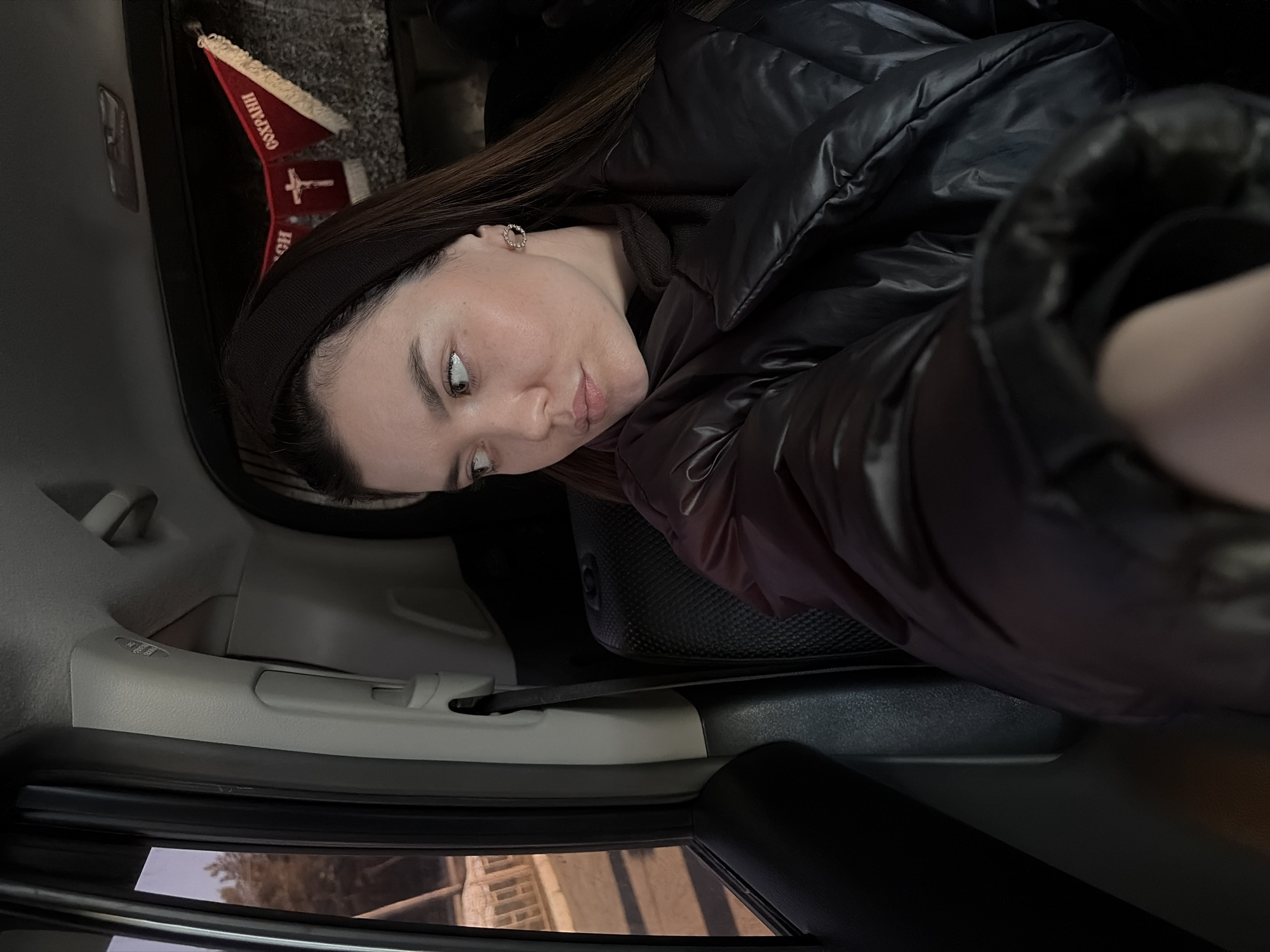 A woman is seated in a car, likely in the back seat. She is wearing a black headband and a black, puffy jacket. A decorative item with the Cyrillic words "Сохрани и" (translated to "Save and") and a cross is visible near the top of the frame. The car's interior is predominantly gray and black. The window shows a glimpse of an exterior scene, possibly an urban environment. The location is Chaikovsky, Russia.