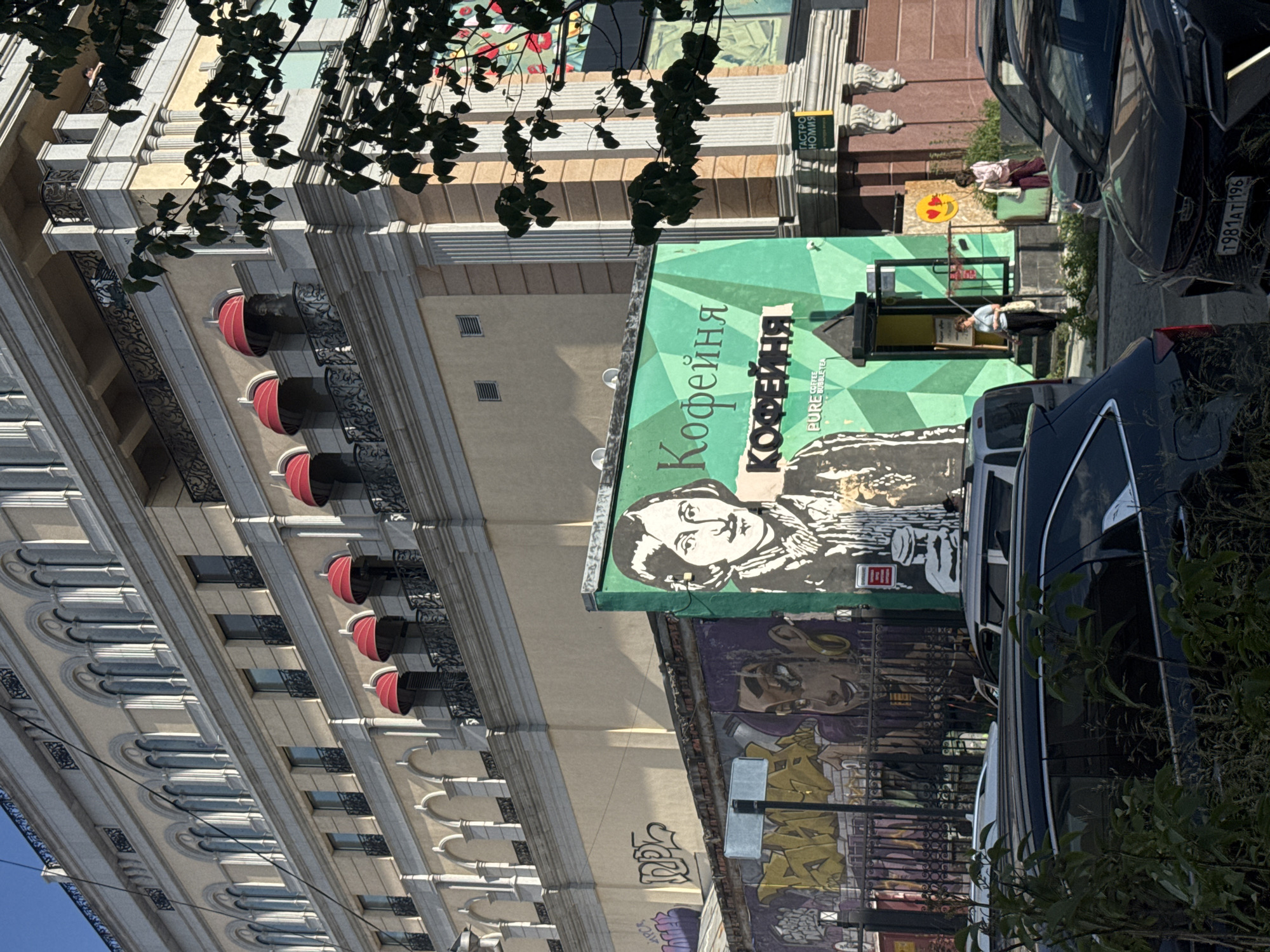 A street view in Yekaterinburg, Russia, shows a building facade with intricate architectural details, featuring arched windows and ornate balconies with red awnings. Overhanging tree branches partially obscure the upper sections of the building. Adjacent to this structure, a vibrant green building displays a large mural of a woman's face, rendered in black and white. Below this, another mural features graffiti art in purple and yellow.
In front of the green building, a woman stands near an entrance, appearing to be engaged in activity. A sign with Cyrillic text is visible on a yellow background. A second woman is standing on a raised platform, leaning against a green surface. Nearby, a yellow sign with a smiley face emoji and two hearts is mounted on the wall.
Parked cars, including a dark-colored sedan with a visible license plate reading "T984AT 196", occupy the foreground, partially obstructing the view of the buildings. The scene suggests a daytime setting with bright sunlight casting shadows. The overall impression is a blend of historic architecture and contemporary urban art, characteristic of a lively city environment.