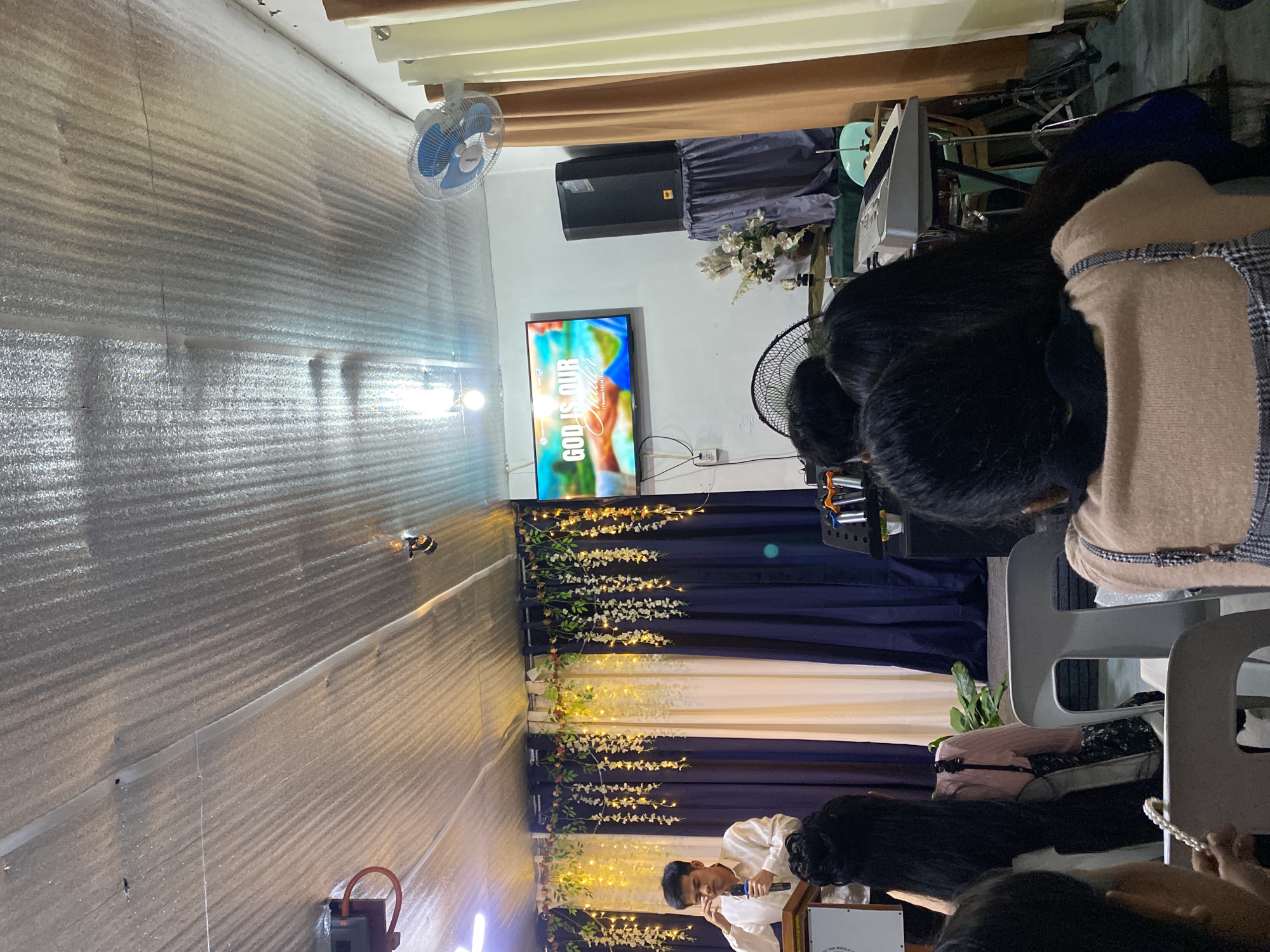 This image captures an indoor religious or community gathering in Davao City, Philippines. A light-skinned male speaker, wearing a light-colored polo shirt, stands behind a light-colored podium, holding a microphone in his left hand while his right hand touches his eye or forehead. The podium's front displays text, partially visible as "THE WHOLE." Behind the speaker, a backdrop consists of dark blue and cream-colored curtains adorned with artificial green vines and yellow string lights.
To the right of the stage, a flat-screen television is mounted on a white wall, displaying the text "GOD IS OUR Healer" with scripture references "2 Chronicles 7:14" and "Isaiah 53:5" over a blurred, colorful background. Below the television, a large black audio speaker rests on a stand draped with a gray cloth, next to a vase with white floral arrangements. Musical instruments, including a keyboard on a stand and parts of a drum set, are visible to the right foreground of the stage. A microphone stand with multiple clips is also present.
A blue electric wall-mounted fan is visible high on the right wall, and another large black fan is partially obscured by audience members in the lower right. The ceiling consists of corrugated, possibly insulated, silvery panels with visible seams, wiring, and small lights.
Several audience members, primarily identifiable by their dark hair, are seated in gray chairs, facing the speaker. One individual in the immediate foreground holds a white beaded item. The room's overall lighting is moderate, with light sources visible on the ceiling and incorporated into the stage decor. Brown and cream curtains are visible on the far right wall.