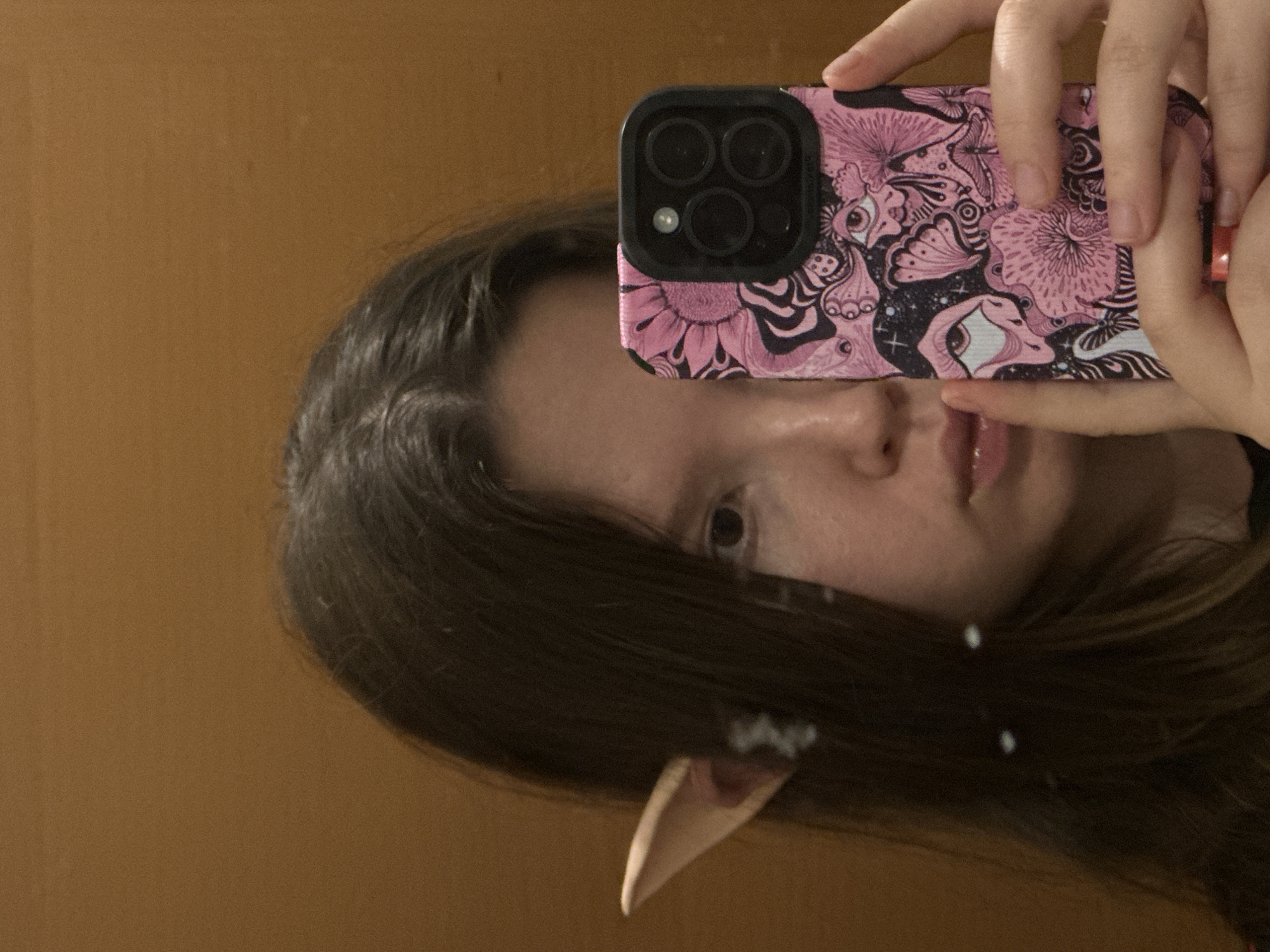 The image captures a mirror selfie, rotated to a vertical orientation, showing the reflection of a person from the chest up. The person has long, dark hair and a neutral expression, with only one eye and part of their face visible. Their left hand holds a smartphone, covering part of their face. A distinctive prosthetic elf ear is visible on the lower left side of the frame, emerging from under the hair.
The smartphone has a black camera module with three lenses and a flash, consistent with a modern iPhone. The phone case is pink with intricate black and white designs, featuring elements like flowers, eyes, butterflies, and other psychedelic or fantasy-inspired patterns. The person's fingers are visible gripping the phone.
The background is a plain, solid brown wall or surface, reflected in the mirror. There is no visible text. The setting appears to be indoors in Budva, Montenegro.