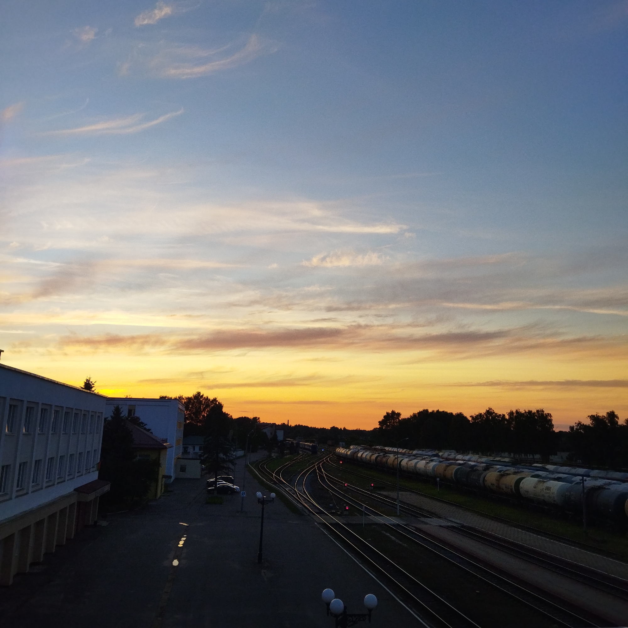 The image captures a train yard in Polatsk, Belarus, during a sunset. The sky displays a gradient of colors, transitioning from pale blue at the top to a vibrant yellow and orange near the horizon, with wispy clouds scattered across. In the foreground, to the left, is a modern, multi-story building with numerous windows. Adjacent to it are dark silhouettes of trees and some parked cars. The central part of the image features multiple train tracks curving through the yard. A long train, consisting of numerous tanker cars, is parked on one of the tracks to the right, its white and grey cylinders reflecting the dim light. The ground appears to be asphalt, with some puddles reflecting the sky's colors. Streetlights with multiple lamps are visible along the tracks, adding to the scene's atmospheric lighting. The overall impression is a serene industrial landscape at dusk.