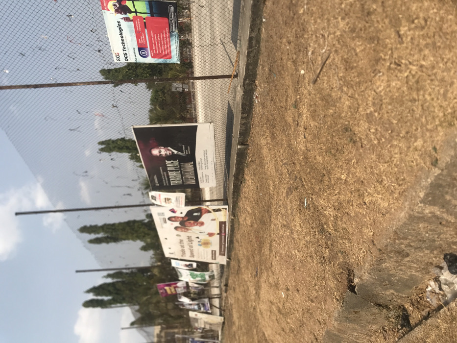 The image captures an outdoor scene in Umudike, Nigeria, during what appears to be a bright, sunny daytime under a clear blue sky with some scattered clouds.
A wire mesh fence runs diagonally across the mid-ground, serving as a display for numerous advertisements and posters. In the foreground, a strip of dry, light brown grass and dirt occupies the lower right section, bordered by a concrete curb or edge to its left.
Several large billboards are prominently attached to the fence. The top-most legible one is red and green, advertising "OGS Technologies" and listing services such as "CCTV," "Networking," "Software Development," "Web Design & Hosting," "Training," and "Sales & Maintenance."
Below this, a significant billboard with a black background features a portrait of a bald man in a suit. Text on this poster reads "THE TREASURE PLACE INTERNATIONAL," followed by details like "TPW Global," "Prayer & Worship," "Every Thursday," "5:00 PM," and a specific location: "RCCG Strong Tower, Umuahia - Aba Rd, Beside Ubakala Market, Umuahia, Abia State, Nigeria."
Further along the fence, another noticeable billboard with a white background displays several people and advertises "Trade at the Speed of Light," with partial mentions of "Premier League" and "Fantasy Football." Numerous other smaller and indistinct posters are also visible further down the fence line.
Behind the fence, a line of green trees is visible, separating the fence from the clear sky. No specific human activity is depicted, but the prevalence of advertisements suggests commercial promotion in a public area.