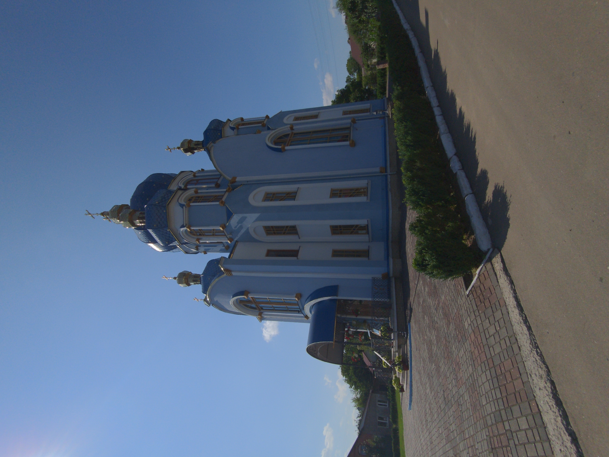 The image depicts a large, light blue church with a blue, golden-domed roof and golden crosses. The church has several windows with arched tops and golden trim. In front of the church is a paved walkway made of red and gray bricks, leading up to an entrance with a blue awning and railings. To the left of the church, there are some houses and trees. In front of the church, there is a neatly trimmed green hedge running along the edge of the paved area, and beyond that, a paved road. The sky is clear blue with a few small white clouds. The sun is shining brightly, casting shadows. The scene appears to be a sunny day in a quiet, possibly rural or suburban, area. The church is the central focus of the image, suggesting it is a significant building in the community. This church is located in the city of Zalishchyky, Ukraine.