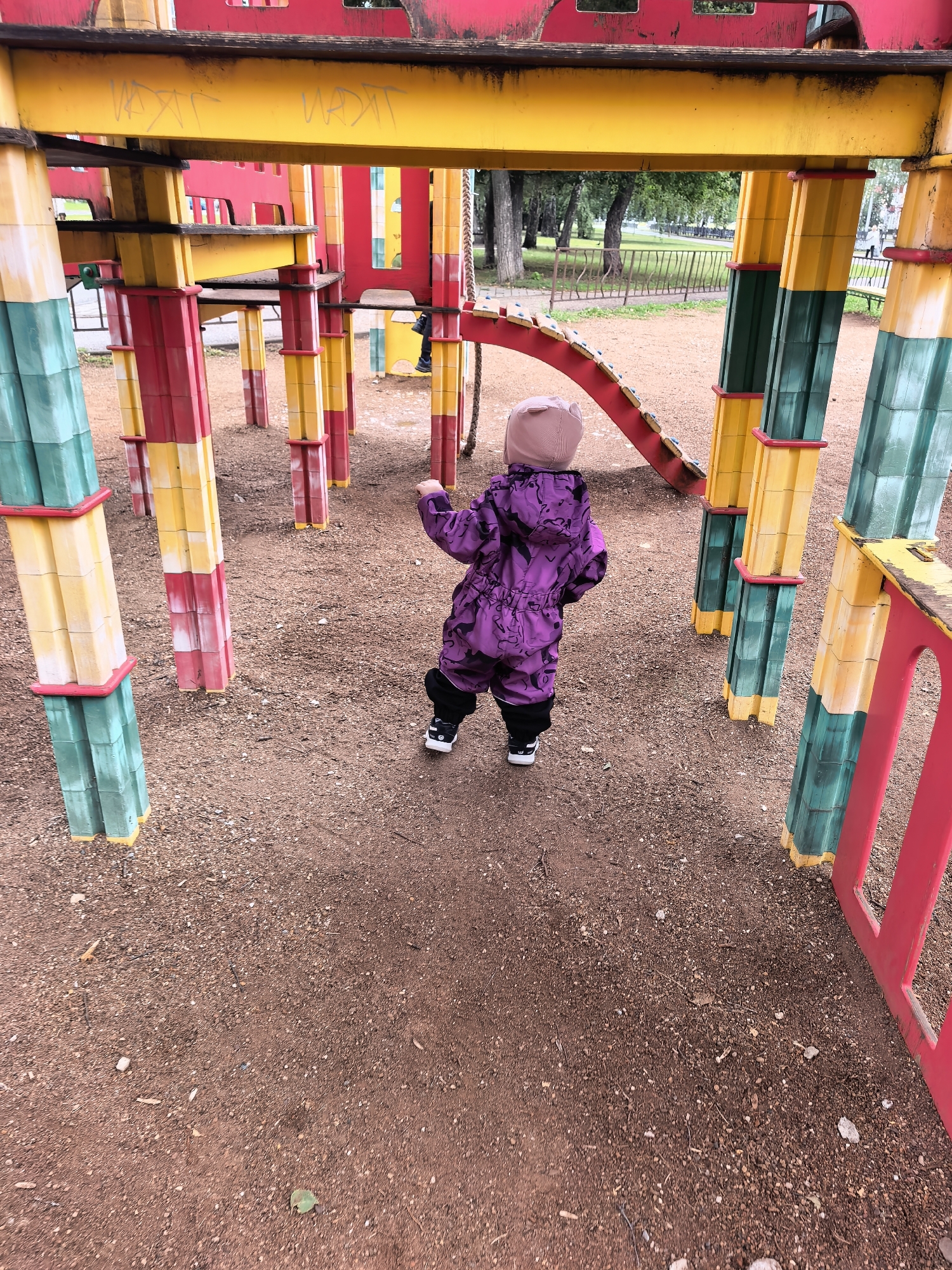 A young child, viewed from behind, is standing on a dirt-covered playground. The child is wearing a purple hooded snowsuit with a black pattern and black shoes. The child appears to be facing away from the camera, toward a playground structure. The playground structure is made of colorful plastic and metal components in yellow, red, and green. There is graffiti on the yellow beam of the structure. Trees and a metal fence are visible in the background, suggesting an outdoor park setting. The ground is covered with fine gravel and dirt. The overall scene depicts a child at a playground in an urban environment.