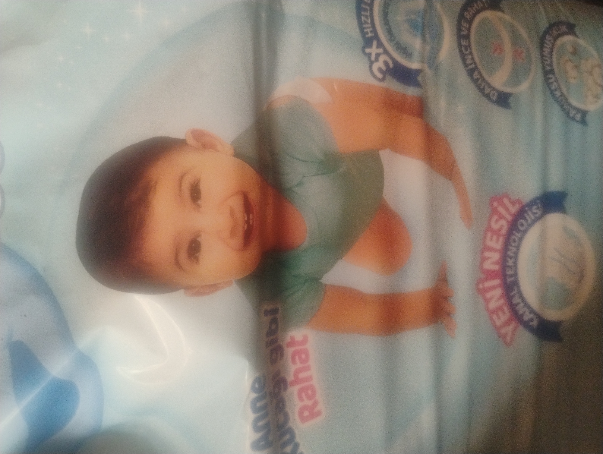 The image displays a close-up of a baby product packaging featuring a smiling baby crawling on a light blue background with subtle star-like patterns. Text on the packaging includes "Anne kucağı gibi Rahat" in Turkish, which translates to "Comfortable like a mother's embrace." Below this, the text "YENİ NESİL KANAL TEKNOLOJİSİ" is visible, meaning "New Generation Channel Technology." Additionally, there are circular badges with text such as "3X HIZLI" (3X Fast) and "DAHA İNCE VE RAHAT" (Thinner and Comfortable), and "PAMUKSU YUMUŞAK" (Cotton Soft). The baby is wearing a light teal colored onesie and appears to be in motion. The lighting is soft, and there is some glare across the image, obscuring certain details. There is no visible indication of the image being taken in the city of Rumyantsevo, Bulgaria.