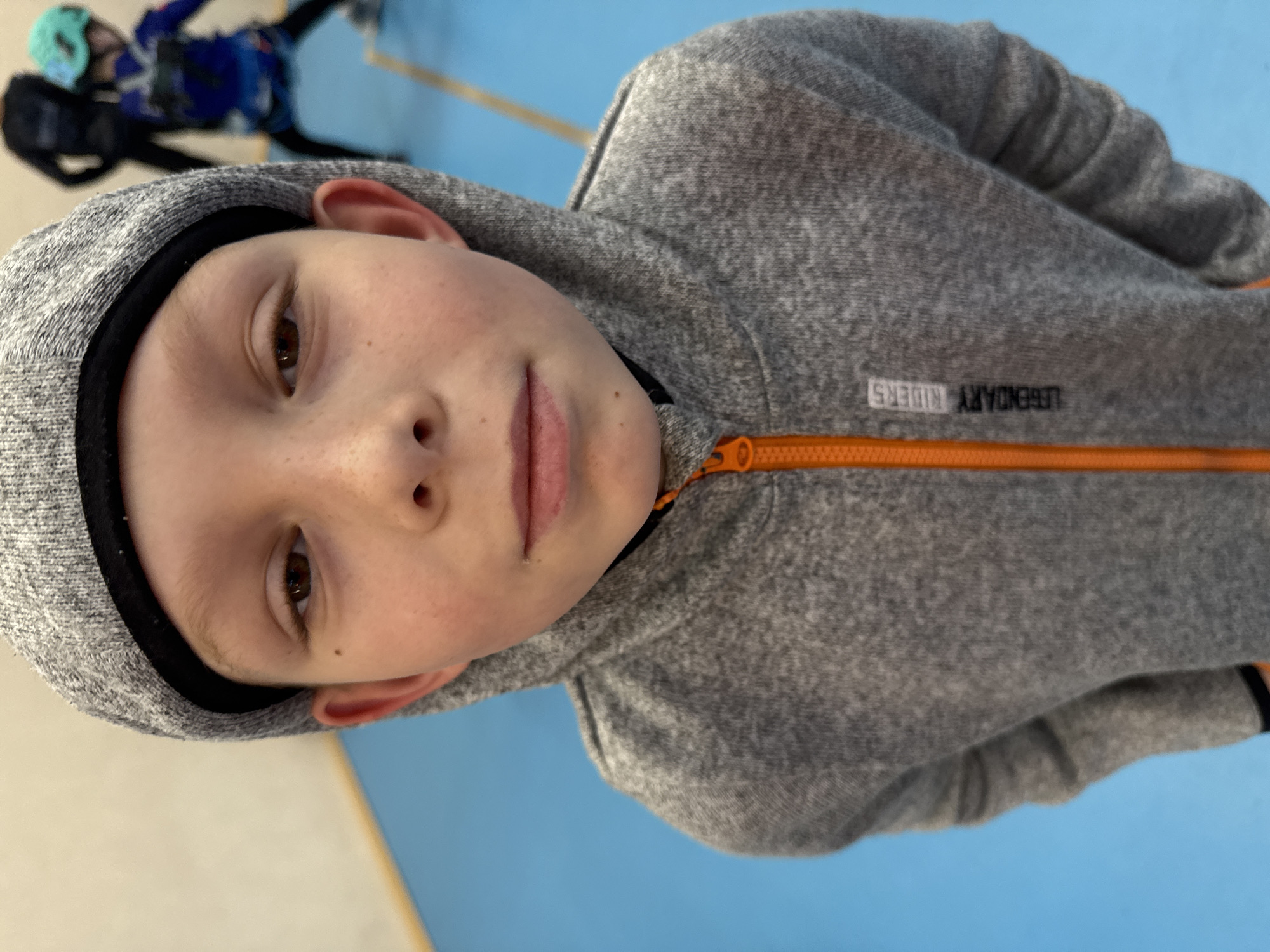 A child with light skin and brown eyes wears a gray hooded sweatshirt with an orange zipper. The sweatshirt has the text "LEGENDARY RIDERS" on the left side. The child is the primary subject of the image, with their face centered in the frame. In the blurred background, another child wearing darker-colored clothing with blue and green accents is visible. The floor is blue, and there's a lighter-colored wall on the left side of the frame. There is no information in this image to determine the location in the city of Berezniki, Russia.
