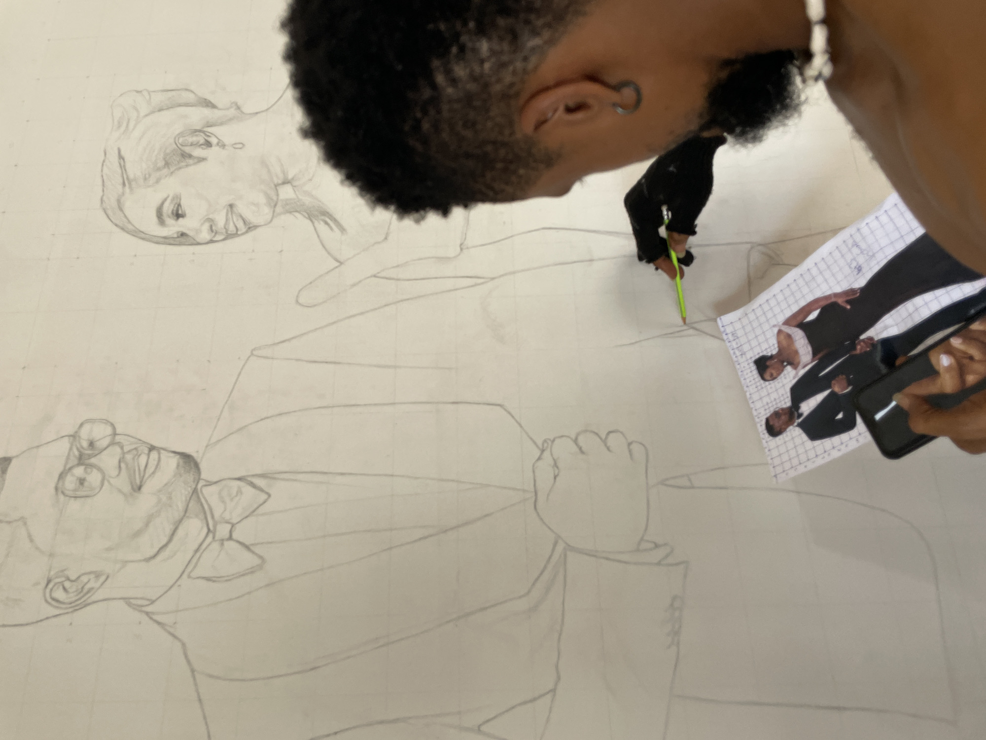 An individual is meticulously drawing a large-scale portrait on a white surface with gridlines. The portrait features two people: a woman at the top and a man below, both wearing formal attire. The artist, positioned above the drawing, uses a green pencil to add detail, referencing a smaller printed photo of the subjects. A smartphone is visible near the printed photo, likely used for reference as well. The environment is indoors, and the focus is on the artistic process. The location is uncertain, but the individual's skin tone suggests the ethnicity of the Ado, Nigeria area.