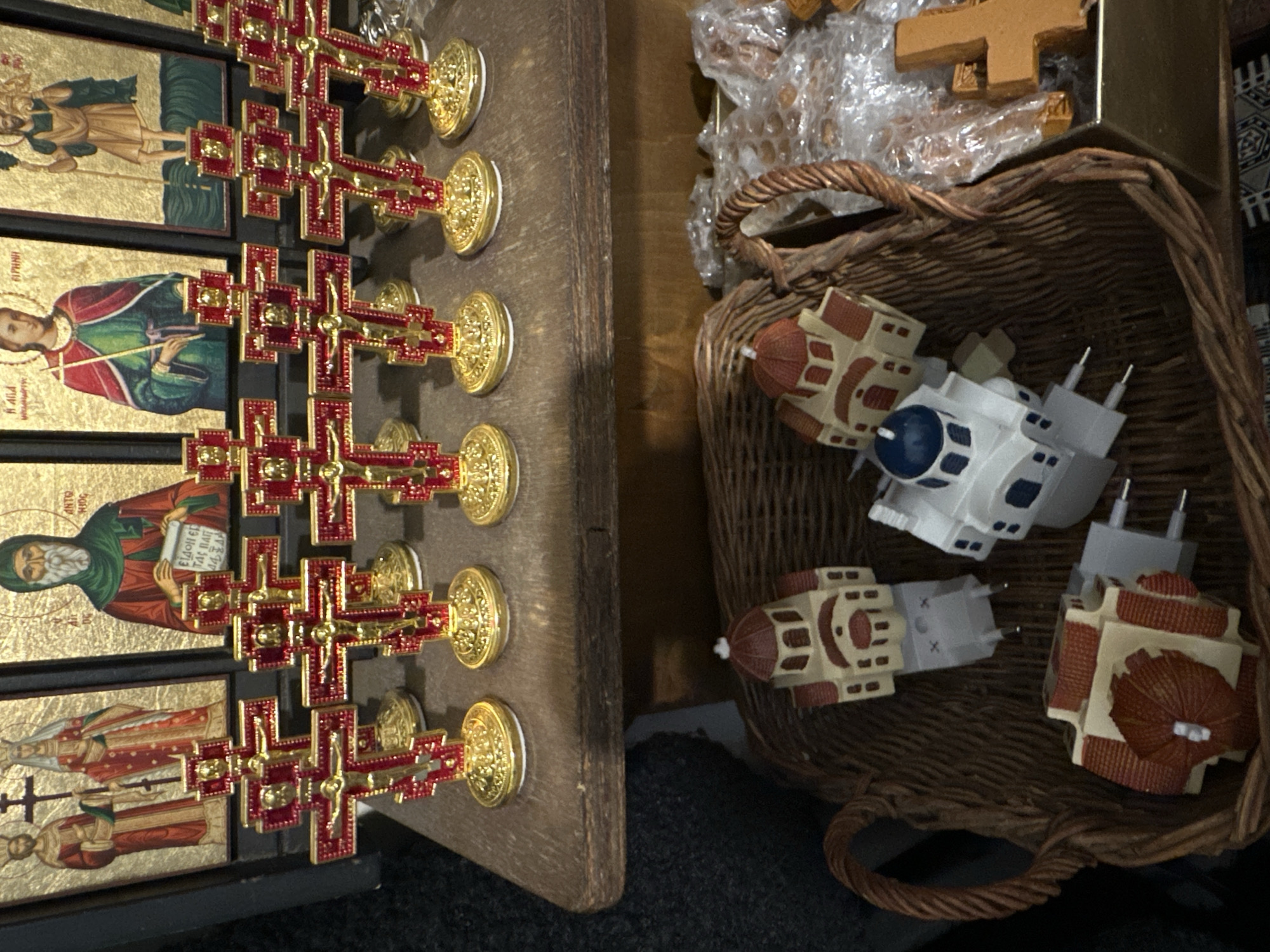 This image displays religious souvenirs arranged on two levels within a retail environment in Maldon, United Kingdom.
On an elevated, dark wooden shelf, eight small, standing crucifixes are arranged in a linear row. Each crucifix features a prominent red cross with golden edges and a golden corpus of Christ. They are individually mounted on ornate, round golden bases. Behind these crucifixes, partially obscured, are multiple gilded religious icons depicting saints in traditional Byzantine or Orthodox style. These icons are set in black frames.
Below the wooden shelf, a brown wicker basket contains additional merchandise. Inside the basket, four miniature building models are visible. Three models are beige with reddish-brown domed roofs, resembling traditional churches, while one is predominantly white with a distinct blue dome, characteristic of Cycladic architecture. Each of these miniature buildings is affixed to a Type C electrical wall plug. To the right side of the basket, other items, including what appears to be a plain wooden cross, are present, some partially wrapped in clear plastic bubble wrap. No people, actions, or direct interactions are depicted.