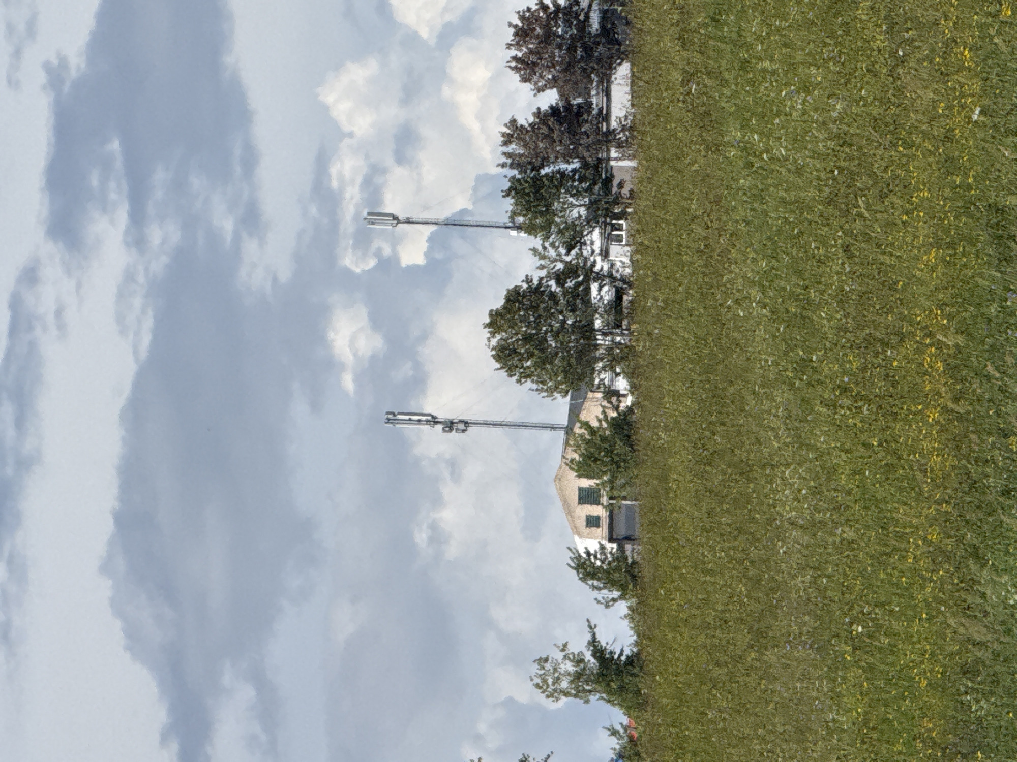 The image captures a rural or semi-rural landscape near Putiniškės, Lithuania, on a day with a dynamic, overcast sky featuring both dark, heavy clouds and brighter, puffy formations. In the foreground, a dense field of tall green grass is dotted with small wildflowers, including yellow and white varieties.
Rising from the middle ground is a low-slung building with a light-colored, possibly weathered, facade. It appears to be an industrial or agricultural structure with a dark, large doorway or opening visible on one side, and several windows, some appearing as dark squares. Adjacent to and behind the building are several trees, varying in size and foliage density. Some trees have dark green leaves, while others, particularly on the right, display reddish-brown foliage, suggesting a change of season or variety.
Dominating the skyline above and behind the building are two telecommunication towers. The tower on the right is more prominently visible, a tall, slender metal structure supporting multiple arrays of antennae at different levels. Another similar tower is visible further to the left, partially obscured by trees. The overall impression is one of a somewhat isolated location, likely on the outskirts of a populated area, characterized by open fields, functional buildings, and modern communication infrastructure. No people are visible in the scene.
