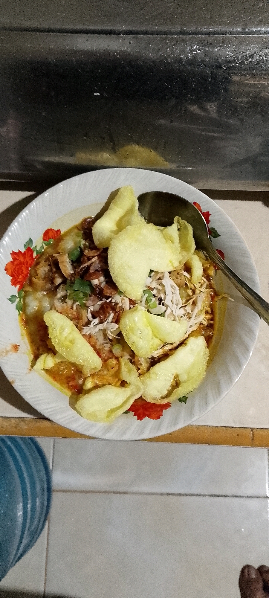 A close-up, top-down view captures a bowl of congee topped with shredded chicken, fried shallots, and light yellow crackers. The congee is served in a white ceramic bowl adorned with a floral pattern of red flowers and green leaves. A metal spoon rests on the right side of the bowl. The bowl sits on a tiled surface. In the background, a dark, metallic surface with a textured appearance is visible. The overall lighting suggests an indoor setting, possibly a kitchen or dining area. This scene is situated in the city of Satriamekar, Indonesia.