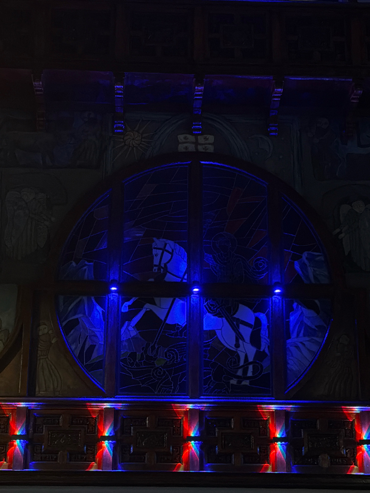The image depicts a circular stained-glass window illuminated by blue lights, located in Batumi, Georgia. The window features a depiction of two horses, one on the left and one on the right, both appearing to be in motion. The horse on the left is white with a black mane and tail, and the horse on the right is also white with a black mane and tail. The stained-glass artwork is framed by dark wood, and the surrounding walls are adorned with relief carvings. Below the window, a decorative wooden panel features carved geometric patterns and is illuminated by colorful lights, casting red, blue, and green reflections onto its surface. The overall scene suggests an interior setting, possibly a church, cathedral, or a similarly ornate building, enhanced by dramatic lighting.