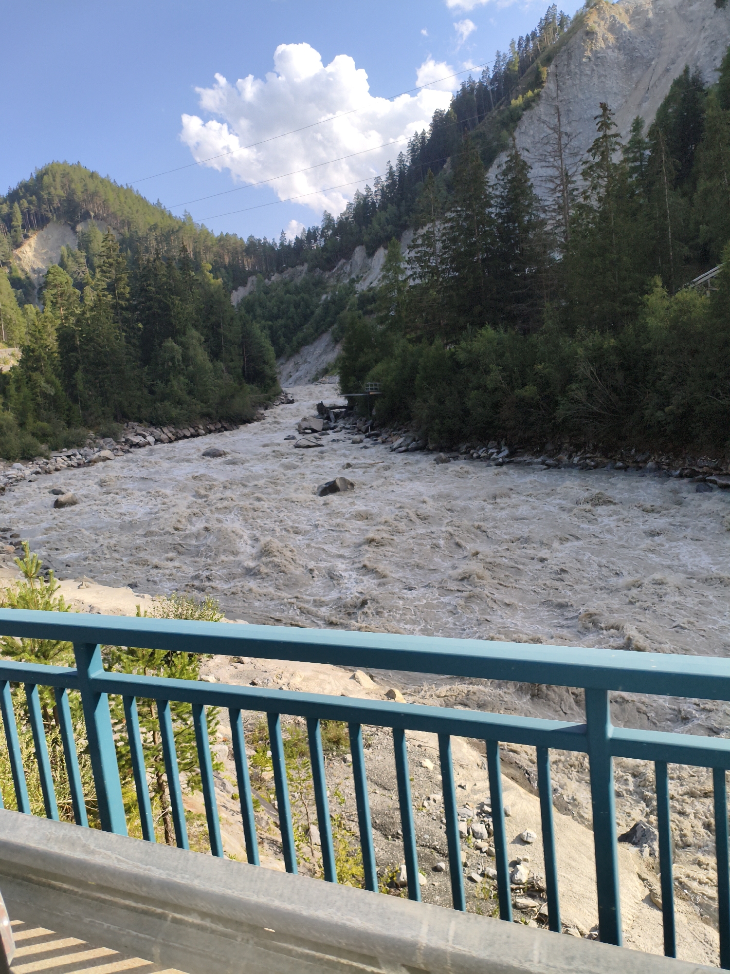 The image shows a wide, fast-flowing river with muddy, turbulent water. The riverbanks are lined with rocks and dense green trees that extend up steep, rocky hillsides. The sky is blue with scattered white clouds. In the foreground, there is a teal-colored metal railing, suggesting the perspective is from a bridge or overlook. The scene appears to be a natural landscape in a mountainous region, likely depicting the Grosser Walserbach river in Winklern, Austria, given its characteristic appearance of a glacial meltwater stream. No people are visible in the image.