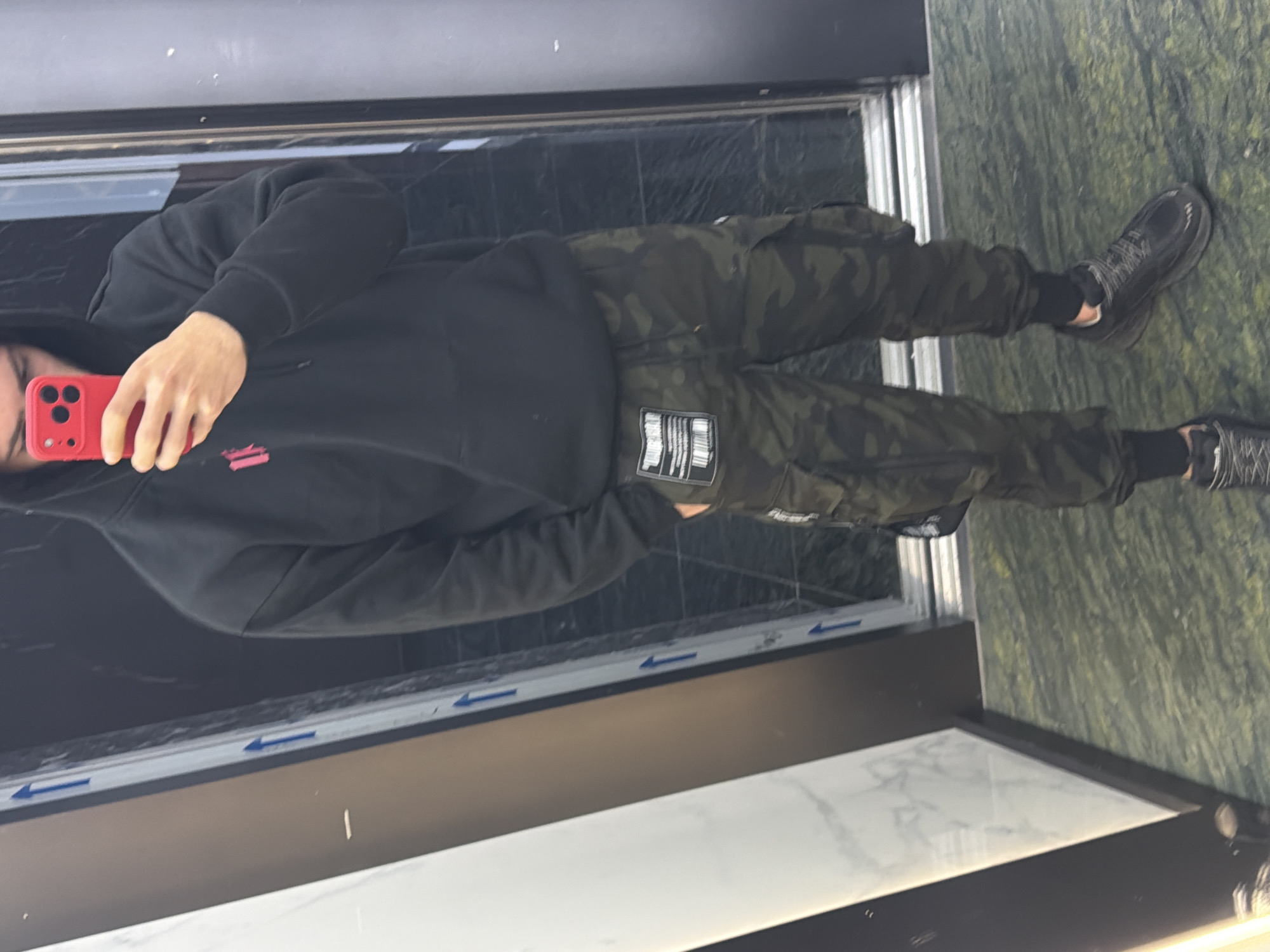 A person wearing a black hoodie and camouflage pants is taking a selfie in front of a window. The person is holding a red phone. The background shows a window frame and a partially visible exterior wall that seems to be made of green stone. The location cannot be determined to be Plovdiv, Bulgaria, based on the image alone.