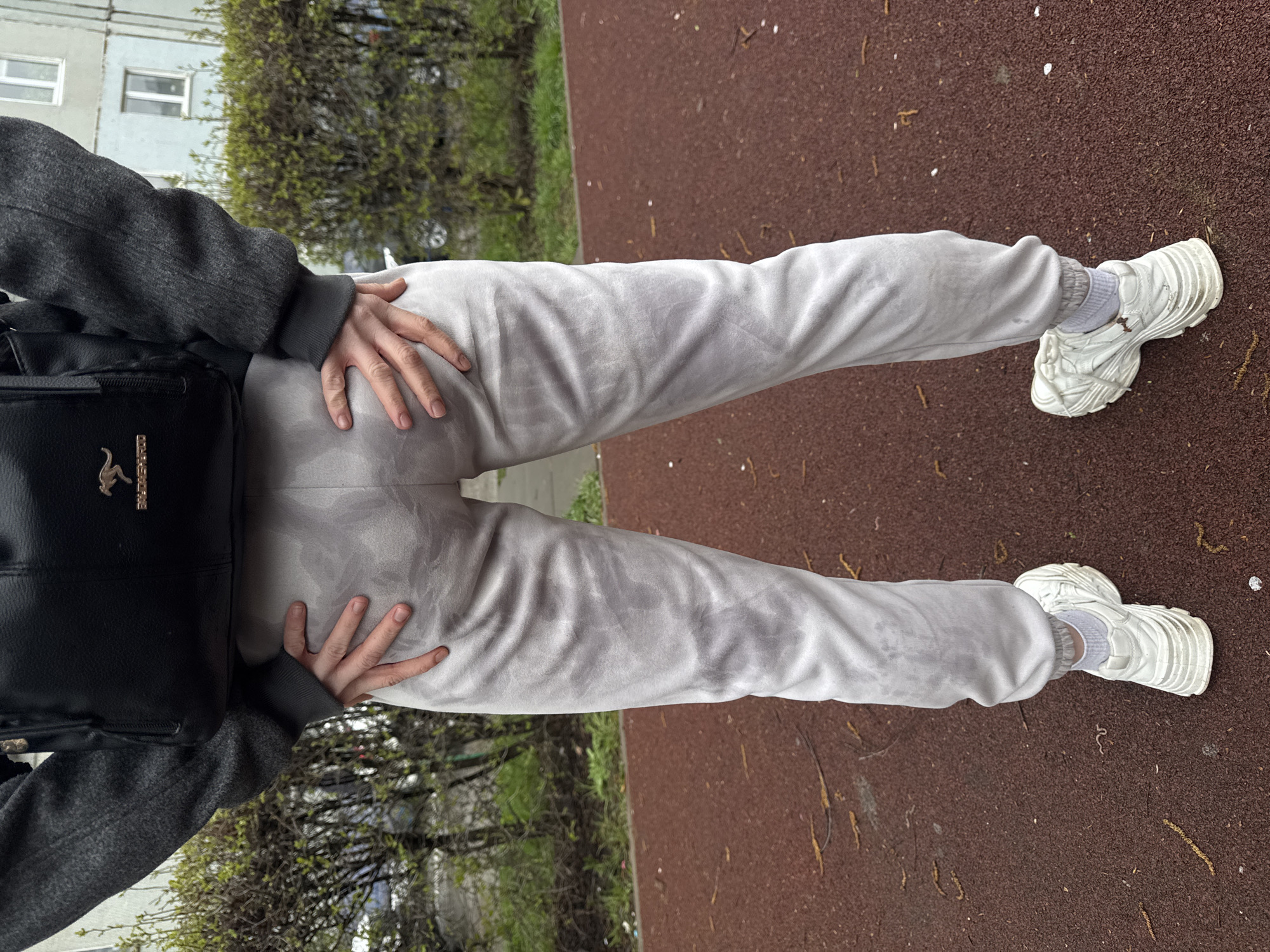 A top-down shot depicts a person standing on a reddish-brown surface. The person is wearing gray sweatpants, white sneakers with visible white socks, and a gray jacket over a black backpack. The person's hands are placed on their hips. The backdrop includes green bushes, trees, and a light-colored building with windows. The location is city of Unknown, Unknown.