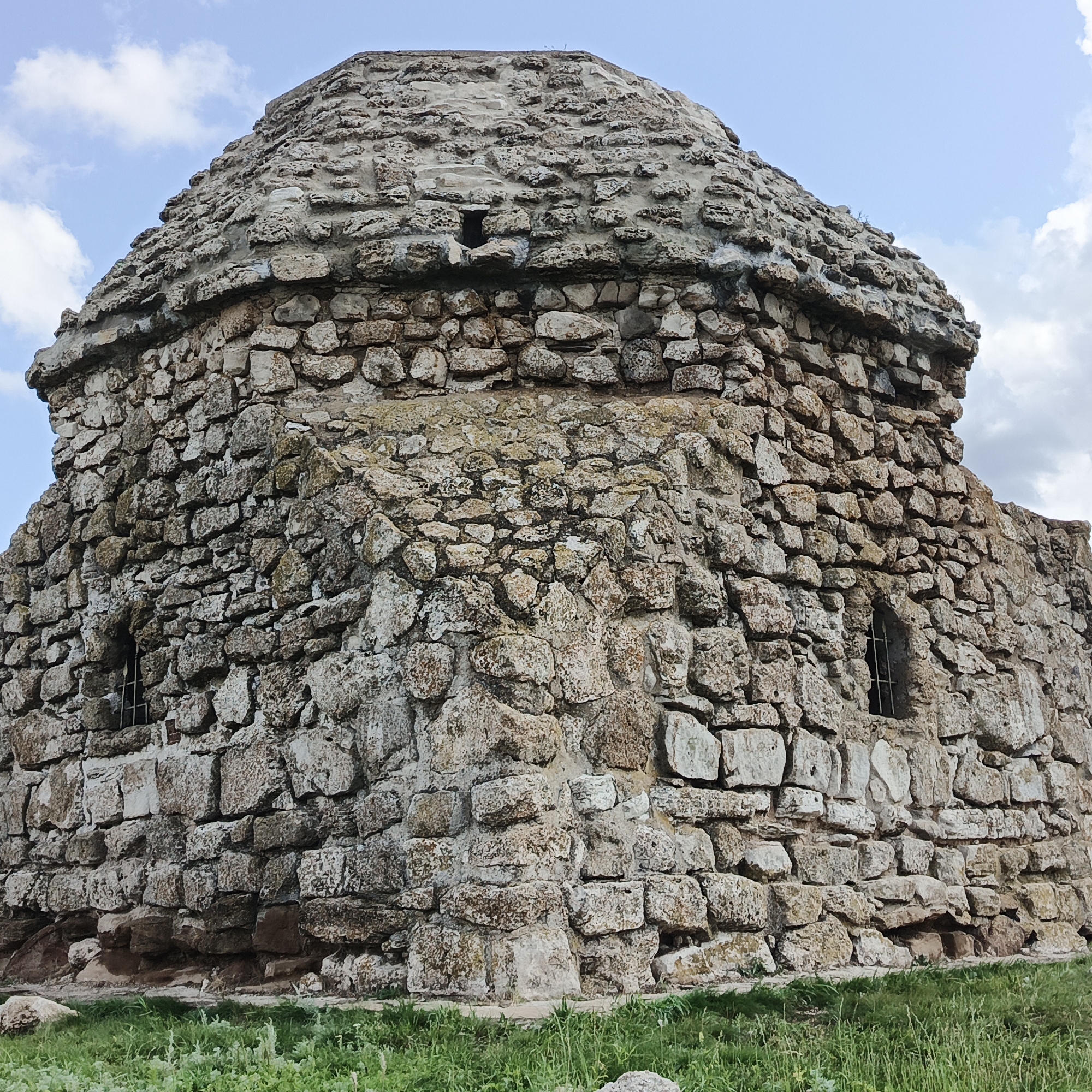 The image shows a stone structure with a domed roof, located in Verkhnie Termy, Russia. The structure is made of rough, irregular stones, with a noticeable dome on top. There are small openings with metal bars on the sides of the structure, resembling windows. The building is surrounded by green grass and is set against a backdrop of a blue sky with scattered clouds. The overall appearance suggests an old, possibly historical, building. There are no people visible in the image.
