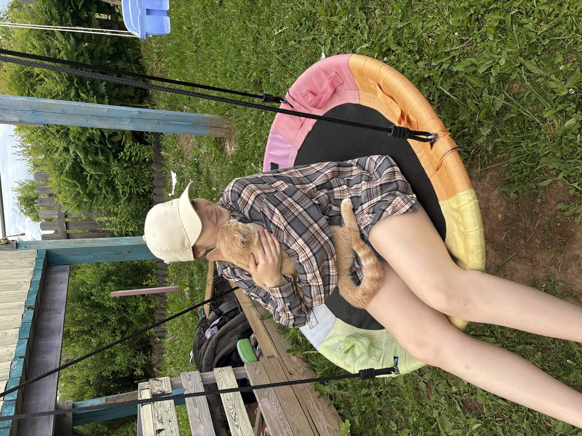 A young Caucasian female wearing a beige baseball cap, a gray and pink plaid long-sleeved shirt, and shorts is sitting on a round, multi-colored outdoor swing. She is holding a ginger cat in her arms. The swing is suspended by black ropes from a wooden structure in a grassy outdoor area. In the background, there is a wooden fence and lush green foliage. The scene appears to be in a rural or suburban backyard setting. The location is identified as Kumyony, Russia.