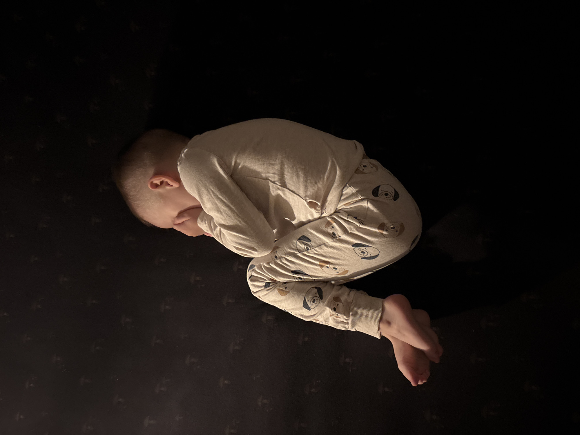 A young child, observed in the city of Chaikovsky, Russia, is depicted lying on their left side in a fetal-like position on a dark surface. The child's head is oriented towards the right, with their hands covering their face. They are wearing light-colored, long-sleeved pajamas patterned with repeating cartoon dog faces. The surface beneath the child is dark, possibly a mattress or blanket, and features subtle, dark grey, repeating mushroom-shaped motifs. The scene is dimly lit, with illumination originating from the upper left, casting a soft glow on the child and parts of the patterned surface.