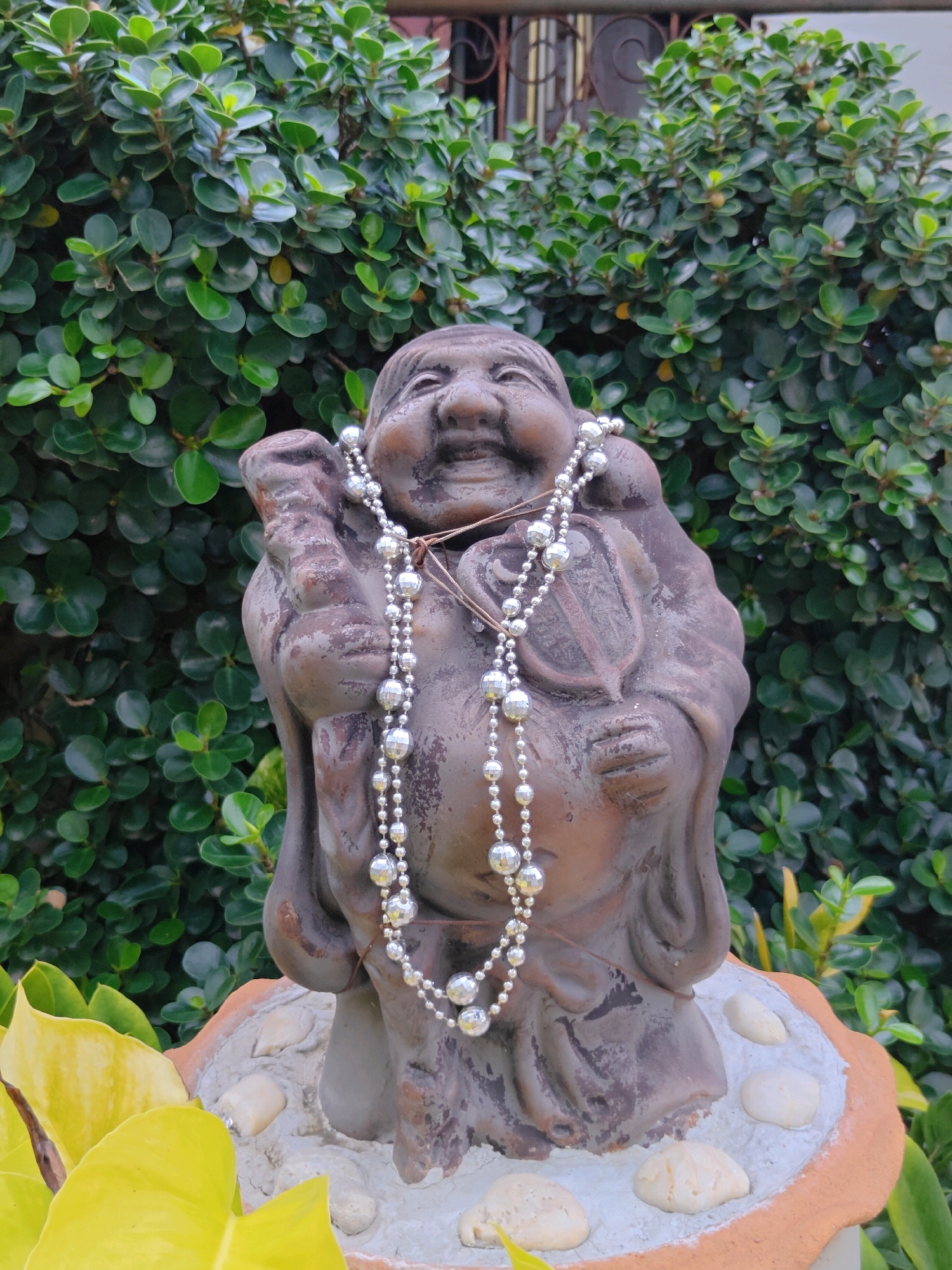 A weathered stone statue of a jovial, plump figure, possibly a representation of Hotei, is adorned with a silver beaded necklace. The statue stands outdoors in front of a dense green hedge. The base of the statue is decorated with pebbles and appears to be made of terracotta. In the foreground, large, bright yellow leaves frame the lower portion of the image. The background includes hints of a building with barred windows and a metal fence. The statue's expression is one of amusement, with its mouth open in a wide smile and eyes crinkled. The sunlight creates highlights on the beads of the necklace and the leaves. The overall impression is one of an outdoor garden setting, possibly in a temple or a residential area within Phra Nakhon Si Ayutthaya City Municipality, Thailand.