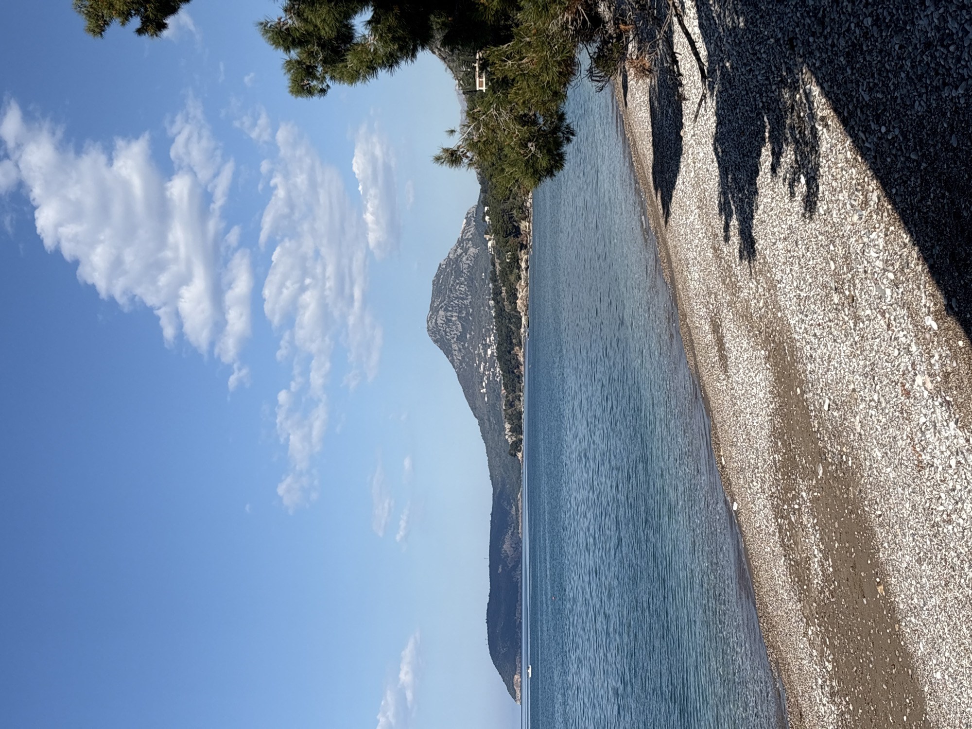 The image shows a beach in Bar, Montenegro, characterized by small, light-colored stones. The Adriatic Sea is visible, with a mountain range rising along the coastline. A partially visible boat is present on the water. The sky is blue with scattered white clouds. Vegetation, including pine trees, is present along the edge of the beach. Shadows from trees are cast onto the pebble beach.