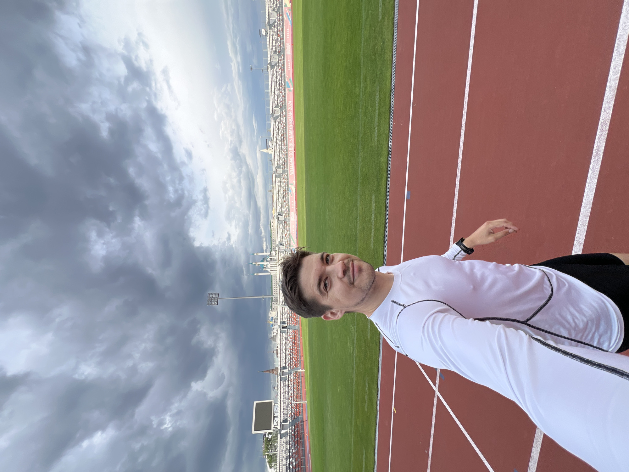 A man in a white long-sleeved shirt and black shorts is taking a selfie on a red running track. Behind him is a green sports field, grandstands, and the skyline of Kazan, Russia under a cloudy sky. The selfie is taken at an angle, showing the man from the waist up.
