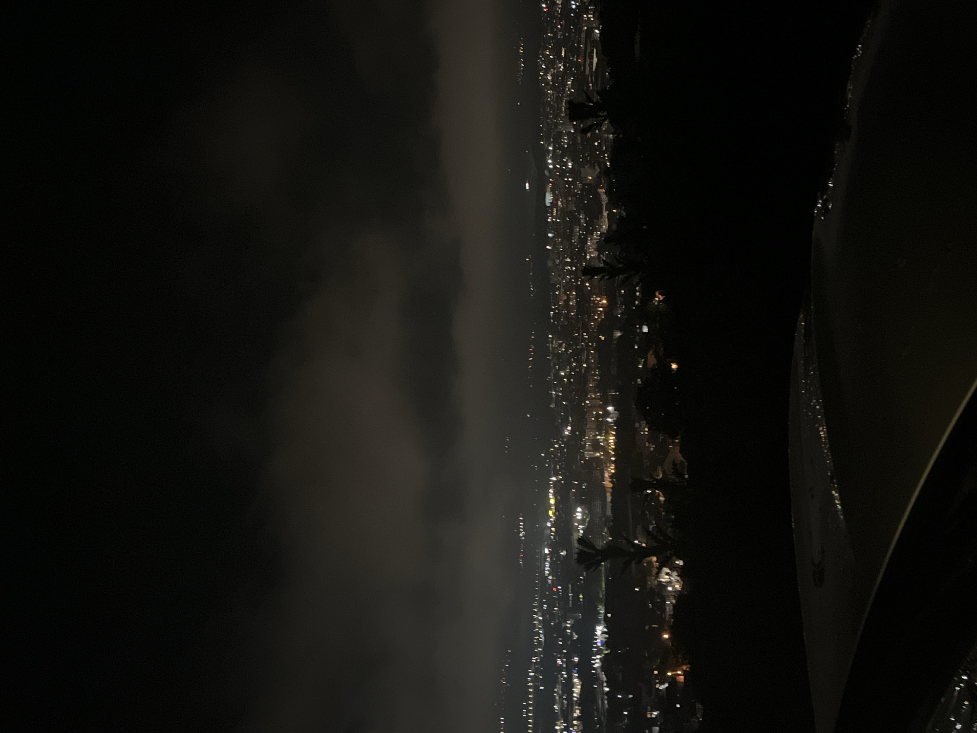 A nighttime shot captures a city lights view, likely taken from an elevated perspective. The city's illumination is partially obscured by a layer of clouds or fog, creating a soft glow in the darker portion of the image. The photograph's composition is oriented vertically, with some parts of an object present on the right edge of the frame. It is difficult to conclusively identify the location as Blagoevgrad, Bulgaria, with the information available.