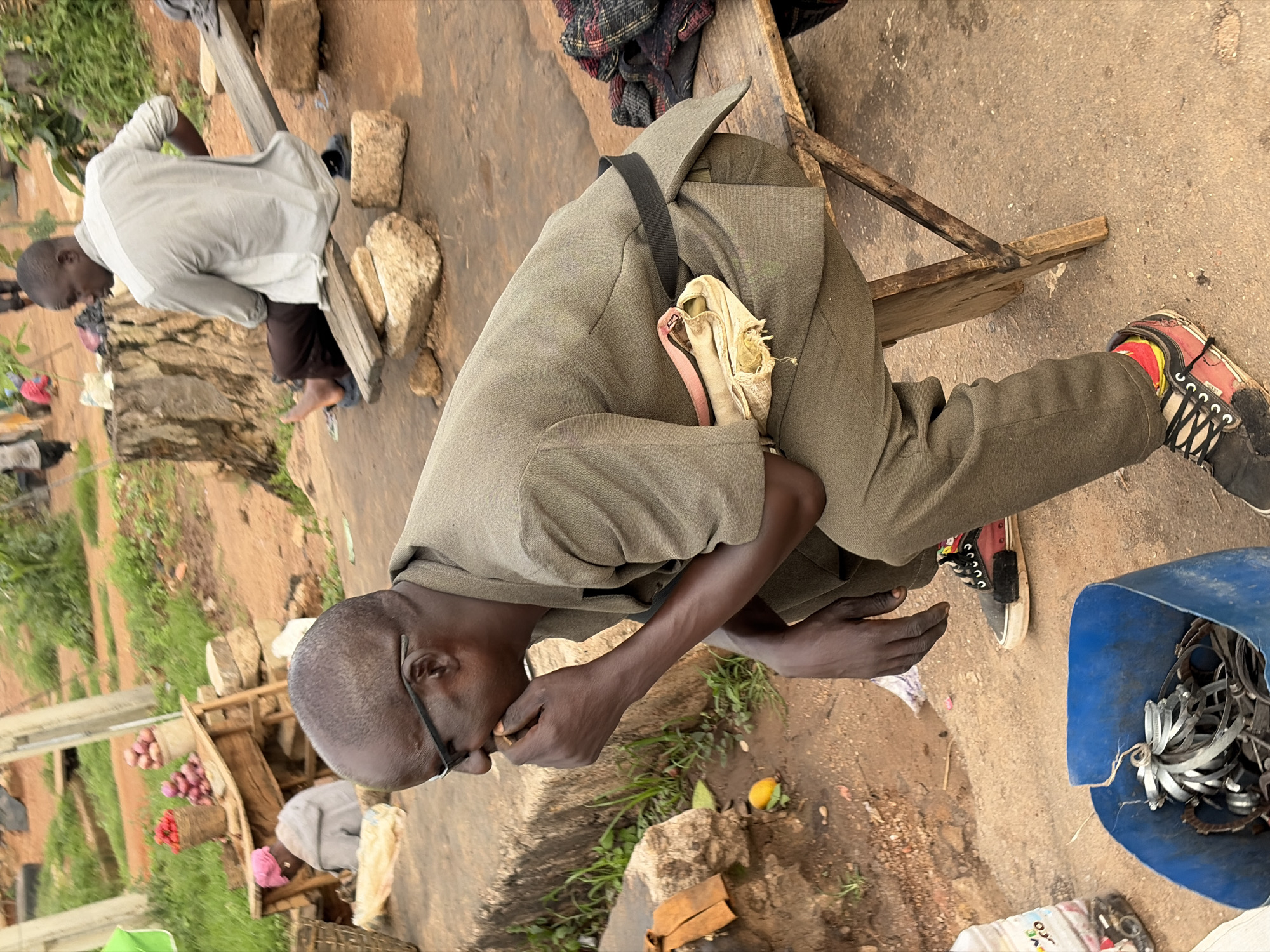 In Gura Top, Nigeria, an outdoor market scene unfolds. In the foreground, a man with glasses sits on a wooden stool, his right hand near his mouth as if eating or contemplating. He wears a khaki suit jacket over a light-colored pouch secured by a black strap around his waist. His trousers are the same khaki color, and he sports multi-colored sneakers. Beside him, a blue plastic bin overflows with metal clamps and other hardware.
In the middle ground, a man in a light gray shirt and dark trousers sits on a makeshift wooden bench supported by stones, facing away from the camera. Further in the background, a stall displays red onions and other produce. Another person, wearing a pink headscarf and gray clothing, is visible behind the stall. The environment is dusty with patches of green grass and vegetation, suggesting a rural or semi-urban setting. A large tree stump is prominent in the mid-background.