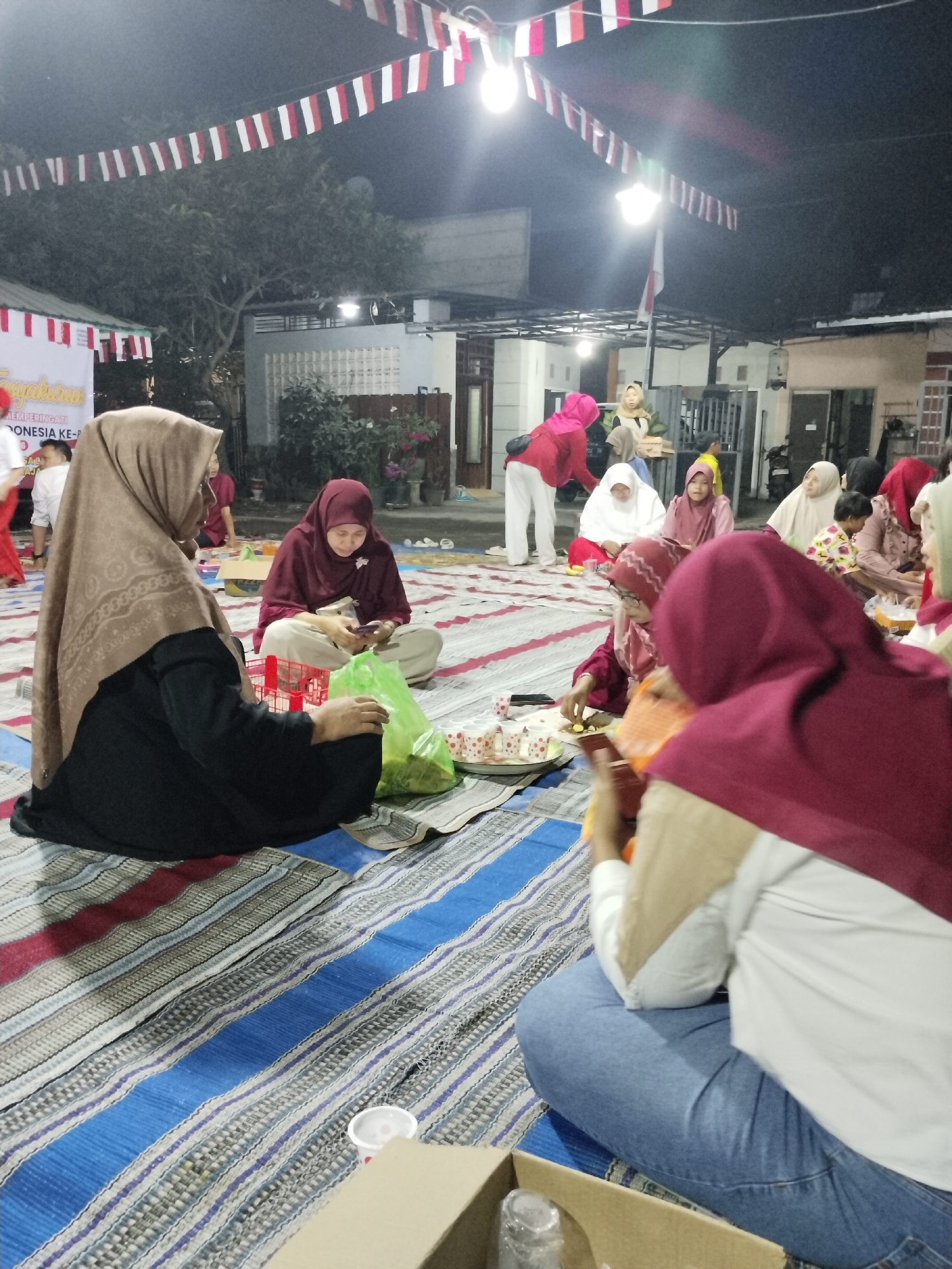 In Lumajang, Indonesia, a group of people, primarily women and children, are gathered outdoors at night, sitting on striped mats. They are celebrating what appears to be a national holiday, as indicated by the red and white Indonesian flags strung across the overhead canopy. The scene is illuminated by artificial lights, casting a warm glow on the gathering. The individuals are dressed in modest attire, with many wearing hijabs. Food and drinks are visible, suggesting a shared meal or a celebratory event. The background shows a residential setting with houses and trees, typical of an Indonesian neighborhood. Some people are engaged in conversation, while others are focused on food or their phones. The overall atmosphere is one of community and festivity.