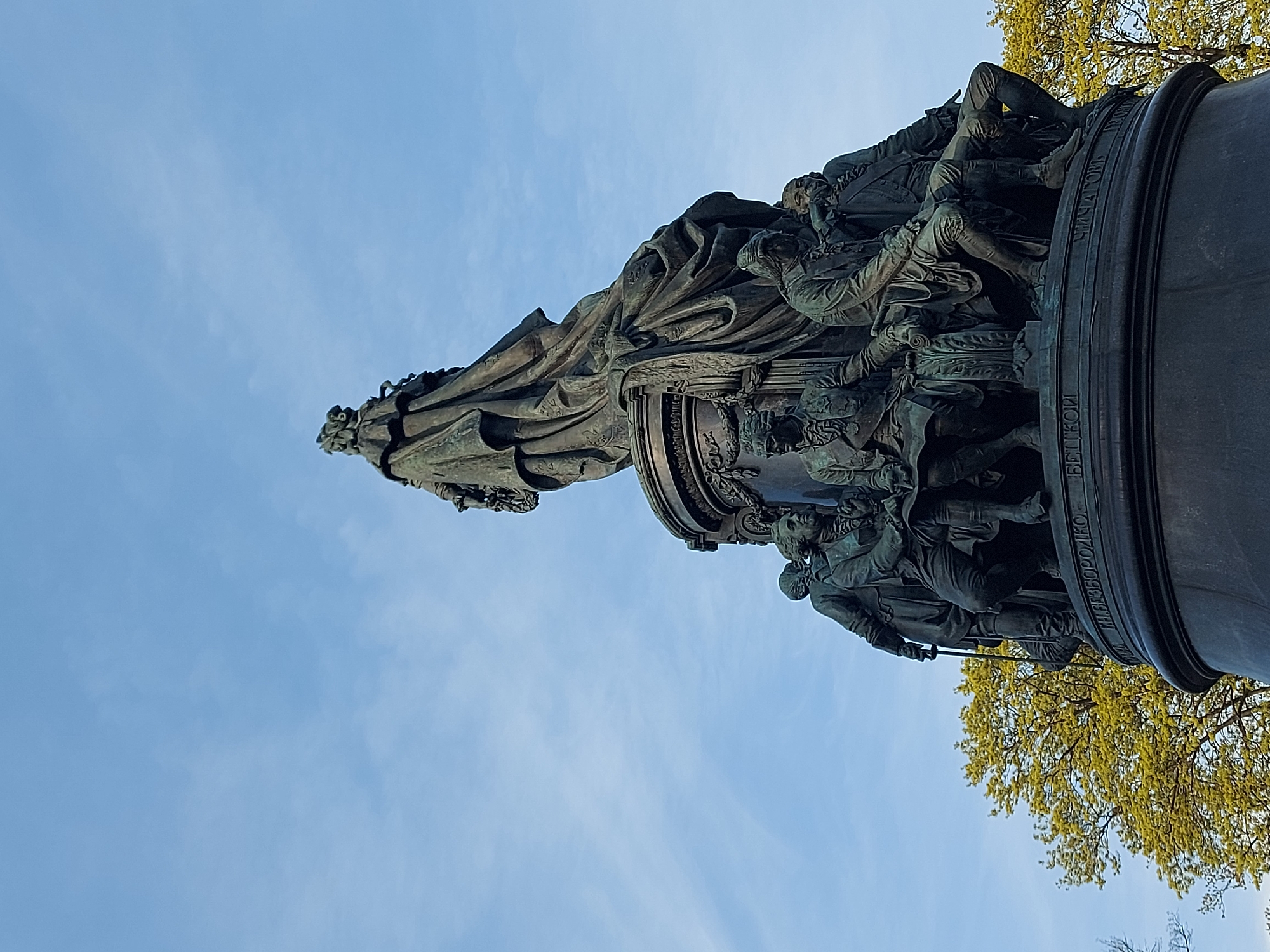 A low-angle view captures the Monument to Catherine the Great in Saint Petersburg, Russia. The bronze statue of the Empress stands atop a cylindrical pedestal adorned with bas-relief sculptures of figures and inscribed text in Cyrillic. A light blue sky, subtly streaked with clouds, forms the backdrop. A tree with yellowing leaves appears in the right portion of the frame.