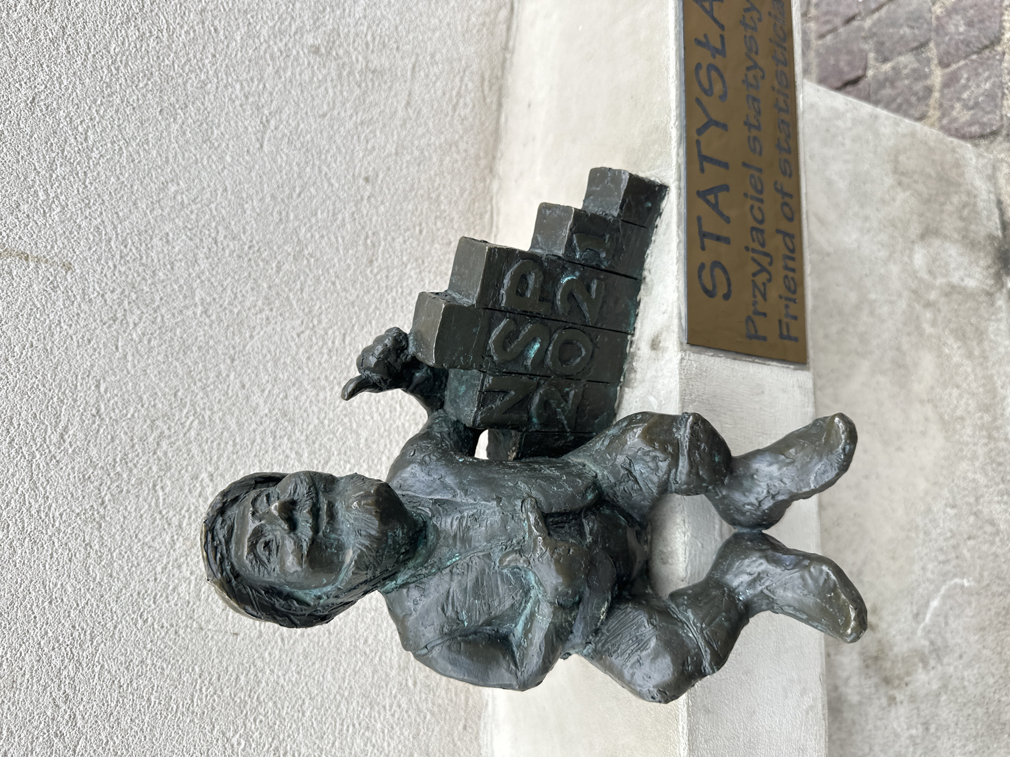 The image shows a bronze dwarf statue, mounted on a stone wall. The dwarf is carrying a stack of blocks on his back with the word "GUS" carved into them. A plaque above the dwarf reads "STATYSTLA Przyjaciel statysty Friend of statistici." This dwarf, known as Statystla (Friend of Statistics), is one of the Wrocław dwarfs located in Wrocław, Poland, and is a monument dedicated to statisticians.
