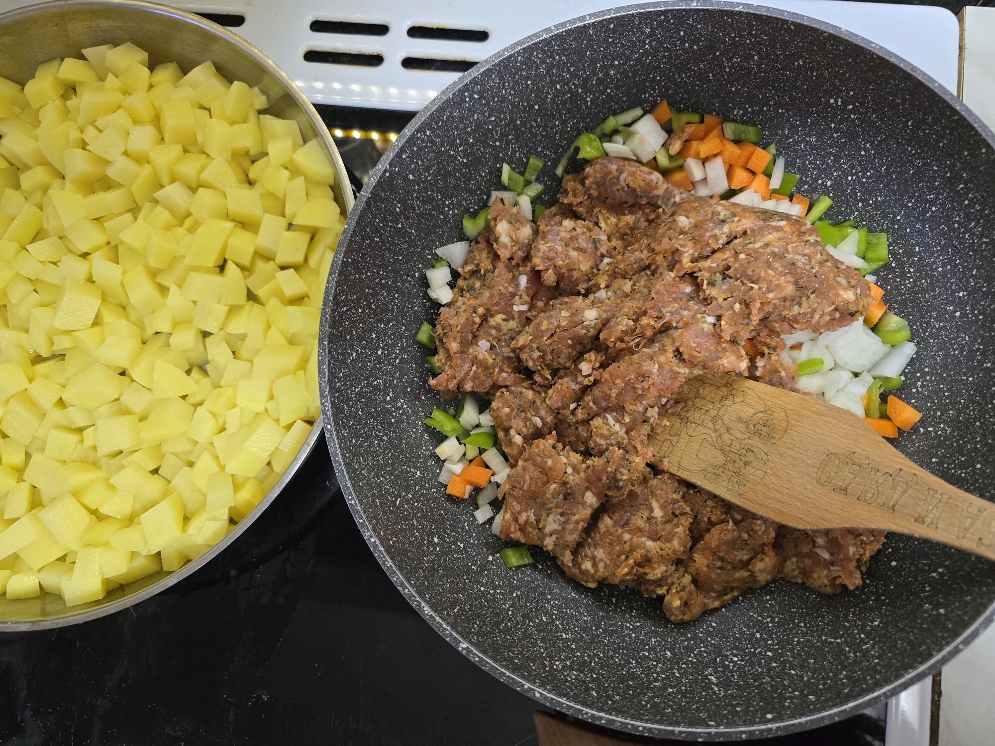 The image shows a culinary scene in a kitchen. On the left is a silver pot filled with diced yellow potatoes. To the right is a speckled gray pan containing raw ground meat mixed with diced green peppers, onions, and carrots. A wooden spatula lies on top of the meat mixture. The setting is likely a domestic kitchen, as evidenced by the stovetop in the background. There is no indication of Burgas, Bulgaria, in the image.