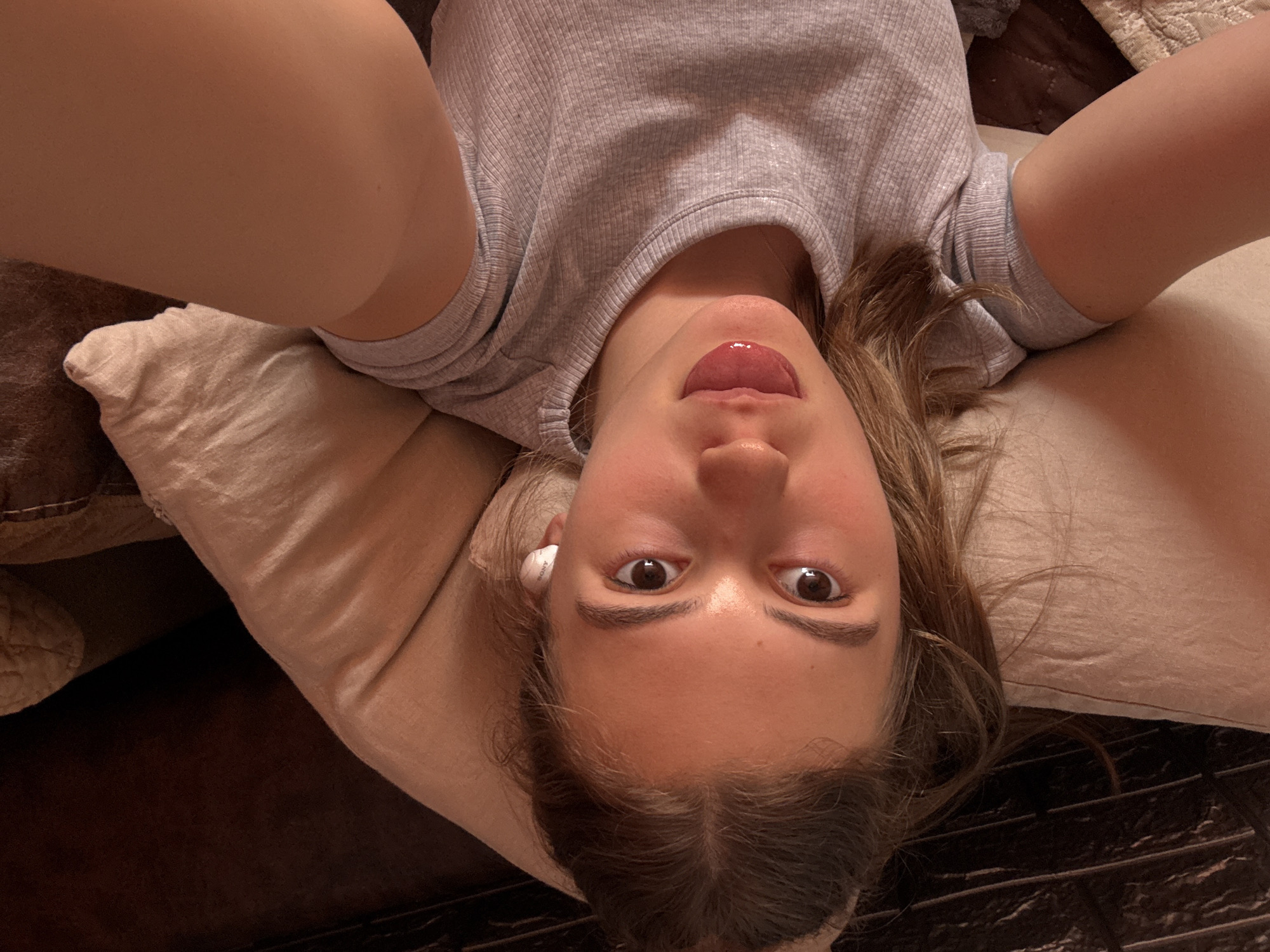 A young woman with long brown hair, a light gray ribbed t-shirt, and a white wireless earbud in her left ear is lying on a pillow on a brown couch. She is holding the camera at an upward angle, displaying an open-mouthed selfie with her tongue partially visible. Her gaze is directed at the camera. This image was not taken in Panagyurishte, Bulgaria.