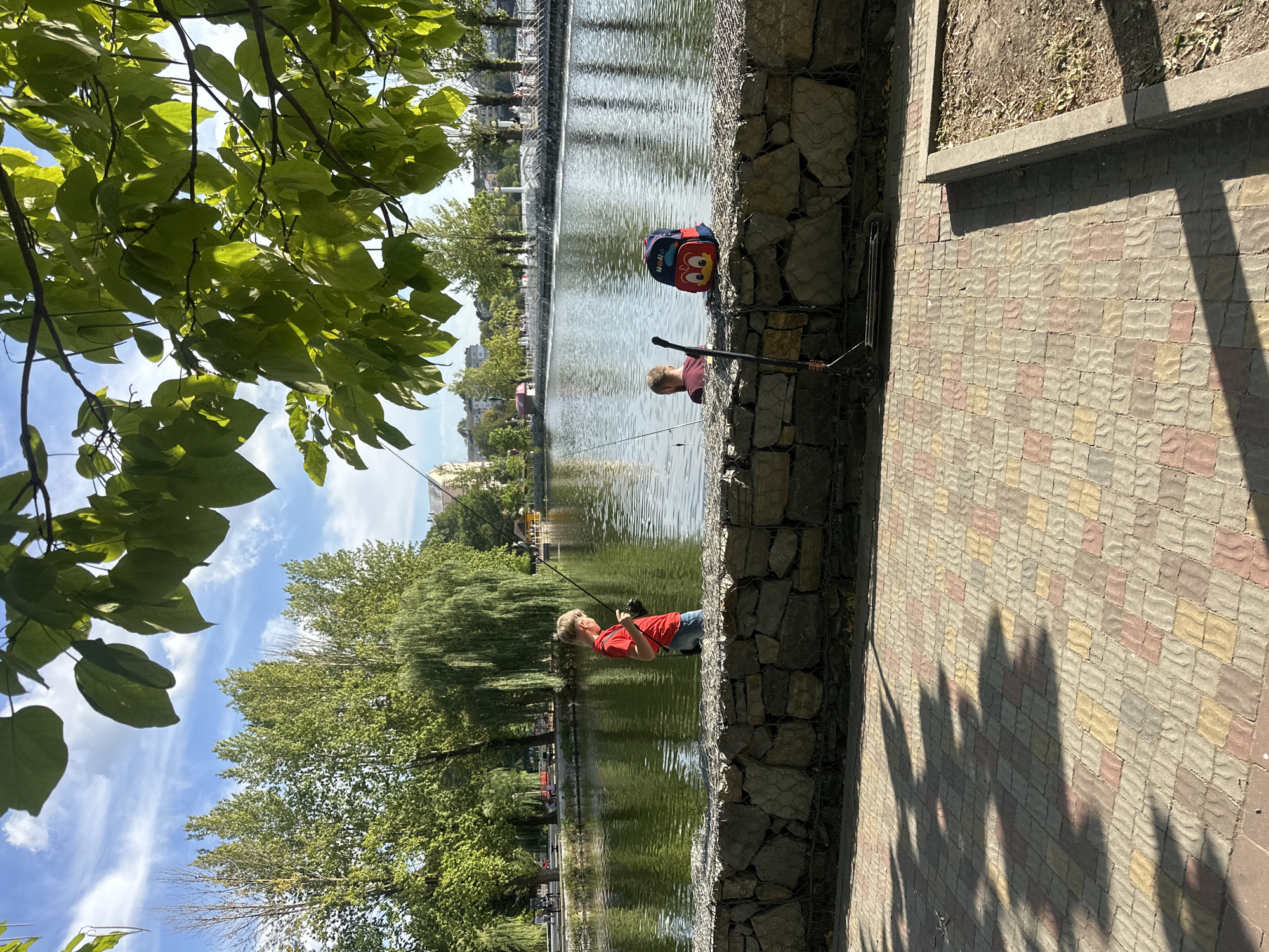 A young boy in a red shirt and blue jeans stands on a stone embankment by a lake in Ternopil, Ukraine, holding a fishing rod. He is looking down at the water. Another person, partially visible, is in the water fishing. A colorful backpack with a cartoon character on it rests on the embankment near the person in the water. A scooter is parked on the paved walkway in front of the embankment. Trees and buildings are visible in the background across the lake, under a partly cloudy blue sky.