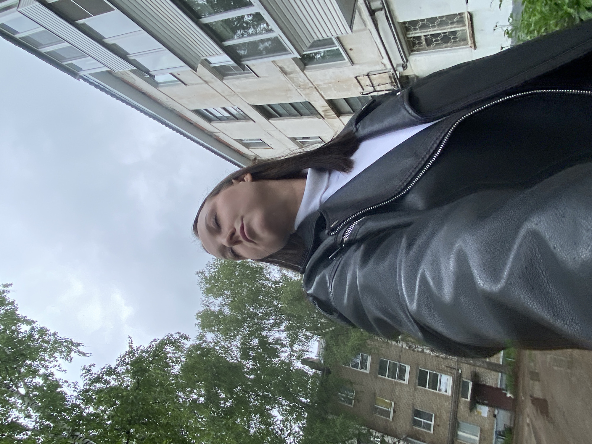A young woman wearing a black leather jacket and a white top stands outdoors. She appears to be taking a selfie from a low angle. The background includes trees, buildings, and a cloudy sky. The buildings appear to be older, possibly Soviet-era apartments. Based on the context provided, it is likely in Chaikovsky, Russia, given that it is where the photographer is from.
