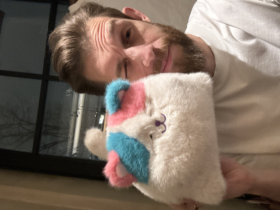 A man winks while holding a white, fluffy, square cat plushie. The plushie features pastel pink and blue stripes on its ears and cheeks, with a purple embroidered nose and closed eyes. The man has short brown hair and a beard and is wearing a white shirt. A window with a dark exterior is visible in the background.