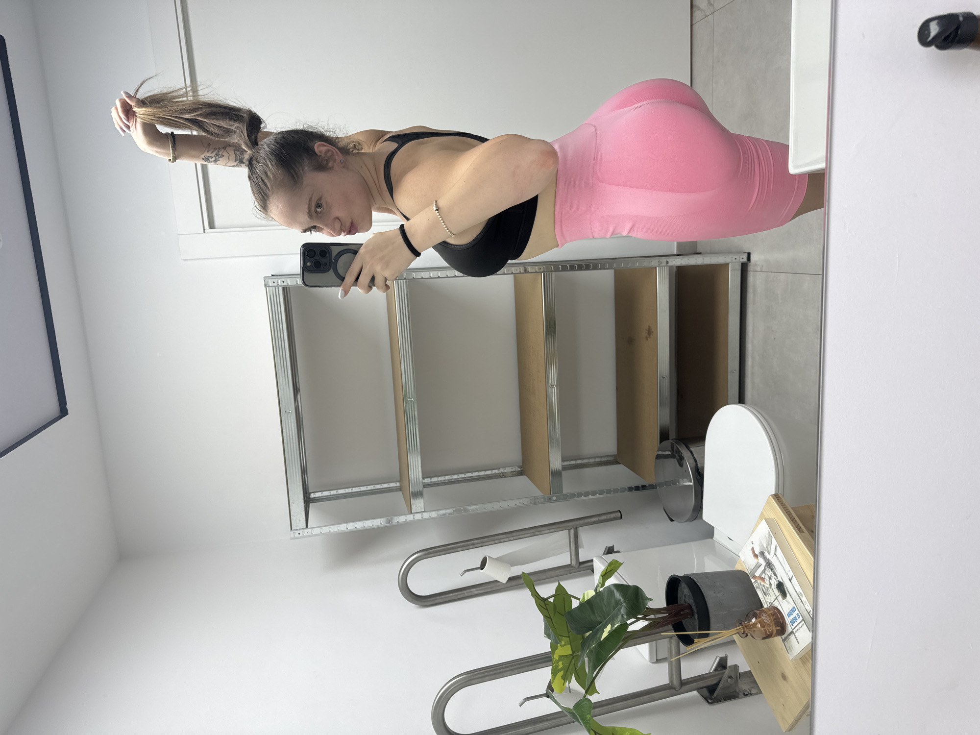 A woman with brown hair, pulled up into a high ponytail, is taking a mirror selfie. She is wearing a black sports bra and pink athletic shorts. She has tattoos on her arms and is wearing bracelets on her wrist. She is in a bathroom, with a toilet, shelves, and a plant visible in the background. The overall lighting is bright and even. There is no indication this image is located in Murcia, Spain.
