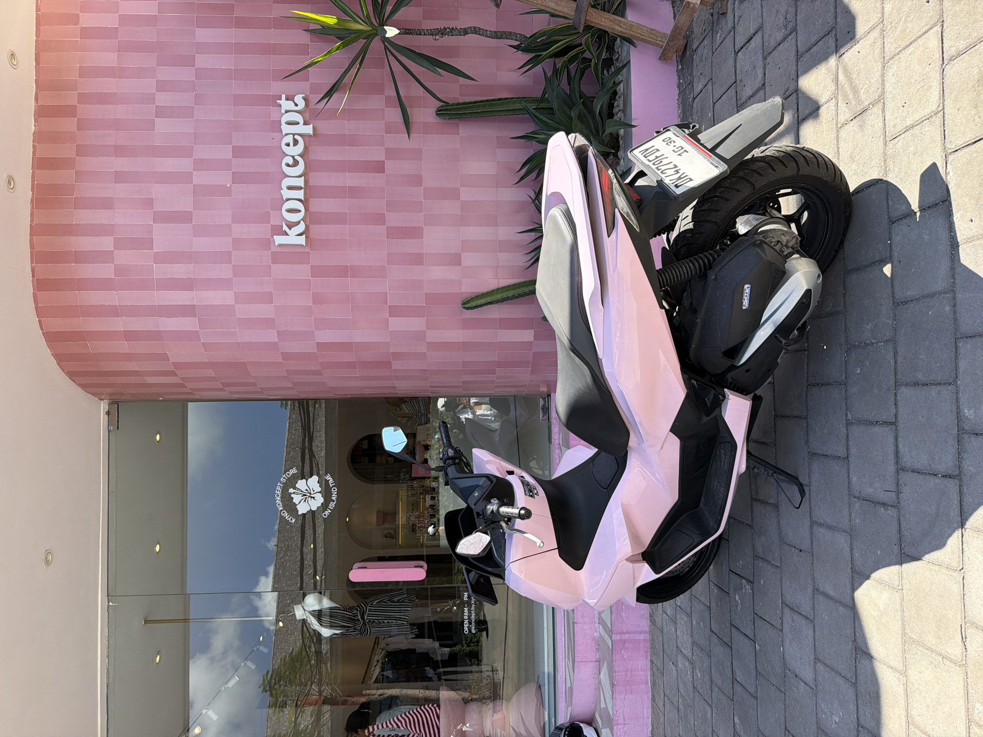 The image captures an outdoor scene featuring a vibrant pink scooter parked in front of a modern store with a pink tiled facade, likely in Kuta Selatan, Indonesia.
The building's exterior is covered in horizontal pink tiles, with the store name "koncept" written vertically in white, raised letters. Adjacent to the building are various green plants, including a tall cactus and leafy, palm-like foliage.
The store entrance features large glass doors and windows that reflect the bright, clear blue sky with some clouds, as well as parts of the surroundings. Text on the glass includes "KYND KONCEPT STORE" and "ON ISLAND TIME" arranged circularly around a flower logo. Other text visible on the glass states "OPEN 9AM - PM" and "@koncept.by.kyr". Through the glass, parts of the store's interior, such as clothing displays and light fixtures, are faintly visible, along with reflections of people, suggesting activity inside.
A pink scooter with black seat and trim is parked on a grey paved sidewalk. Its license plate, "DK4279FDY," is clearly visible, along with the numbers "10-30." The bright sunlight creates strong, distinct shadows of the scooter and nearby objects on the sidewalk, indicating it is daytime, likely mid-day or afternoon. The overall scene suggests a stylish commercial area with a tropical ambiance.