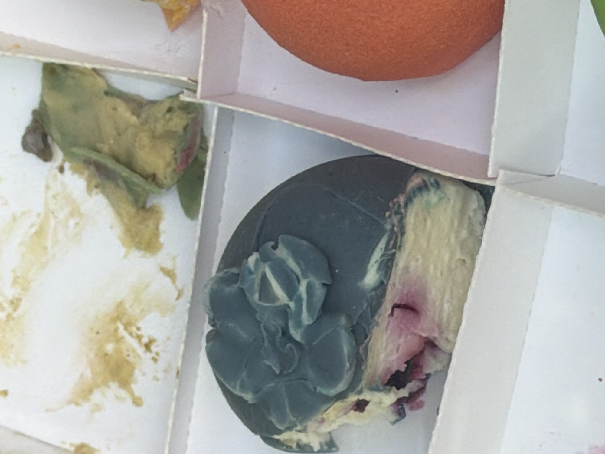 The image shows three compartments of a white cardboard food container. The top right compartment contains an orange, round object. The bottom center compartment contains a dark gray, round object with blue-green mold growing on one section. The object appears to have been cut, revealing a beige and pink interior. The left compartment contains a green and yellow mass with dark brown spots and some green leaves. There are also brown stains on the white cardboard. There is no information provided about the location being in Bishkek City, Kyrgyzstan.