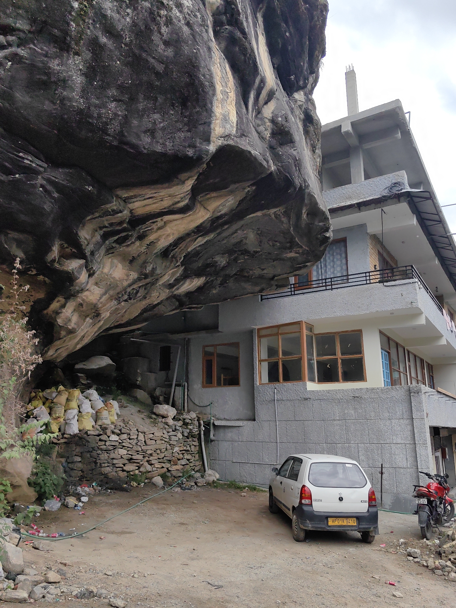 In the city of Vashisht, India, a building is under construction adjacent to a large rock overhang. The overhang casts a shadow over a portion of the building and the ground below. A white hatchback car is parked in front of the building, and a red motorcycle is parked to the right of the car. Bags of construction material, including cement sacks, are piled near the rock face, and a stone wall has been constructed in front of them. Scattered debris litters the ground. The building appears to have multiple stories, with balconies and windows visible. The sky is overcast.