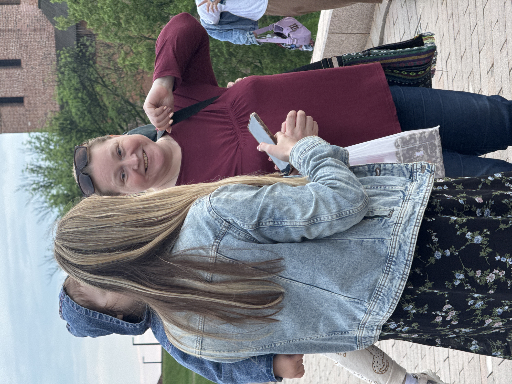 In Nizhny Novgorod, Russia, a fair-skinned woman with short hair and sunglasses on her head smiles, holding a cell phone. She wears a maroon v-neck shirt and a black cross-body bag. A younger person, presumably a girl, wearing a denim jacket and a black floral dress, stands beside her, also holding a cell phone. The scene is outdoors on a paved surface, with greenery and buildings in the background. A bag and other items are visible at the top of the frame.