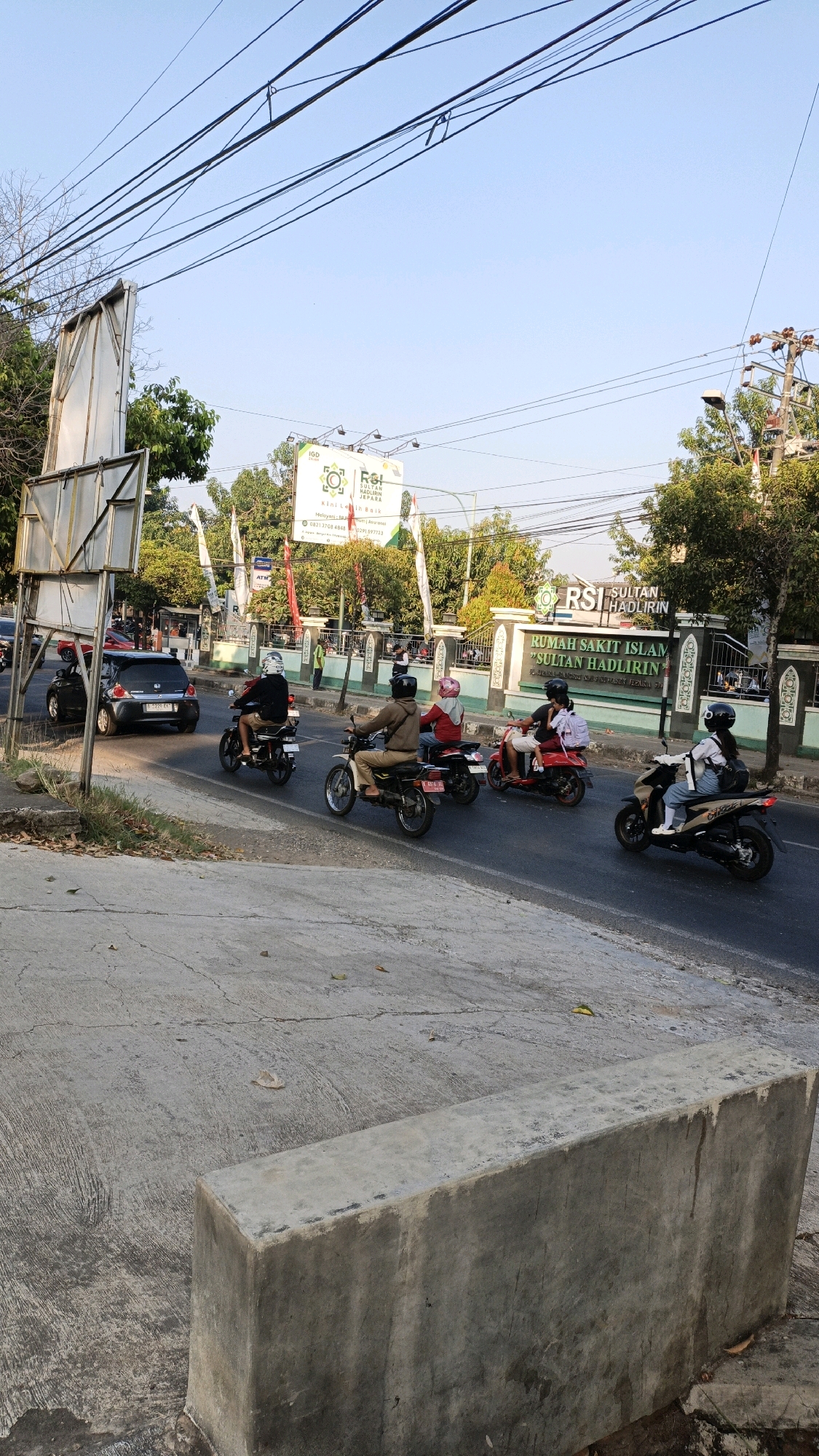 The image depicts a street scene in Kuwasen, Indonesia, with a clear blue sky overhead. In the foreground, a concrete barrier runs along the edge of the road. The road itself is asphalt, with visible cracks and some scattered leaves. Several motorcycles are in motion on the road, carrying riders. One motorcycle is traveling towards the viewer, with a person wearing a light-colored shirt and helmet. Another motorcycle is moving away from the viewer, carrying two individuals, one in a red top and another in a white top. Further down the road, another motorcycle carries a person in a tan jacket and helmet. To the left, a black car is parked. In the background, trees and buildings are visible, including a sign for "RUMAH SAKIT ISLAM SULTAN HADLIRIN," indicating the presence of a hospital. Utility poles and wires are also present, typical of an urban environment. The overall impression is of a bustling street on a sunny day.