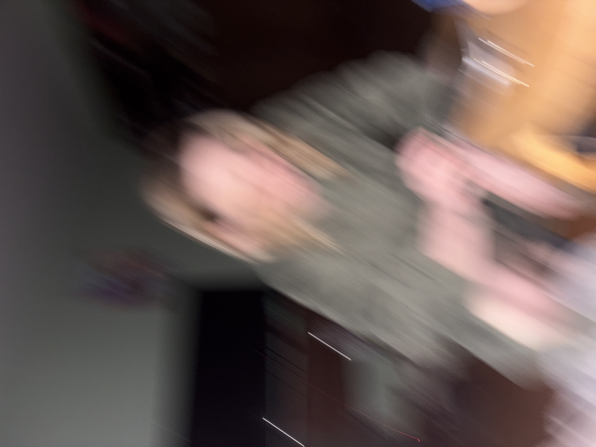 The image is blurry and lacks clear details, making a definitive assessment difficult. However, it appears to depict a person with light skin and blonde hair, possibly wearing a gray garment. The background suggests an indoor environment with a dark area and potentially a wall. Identifying the exact location in Unknown, Unknown is impossible due to the image's poor quality and lack of contextual information.